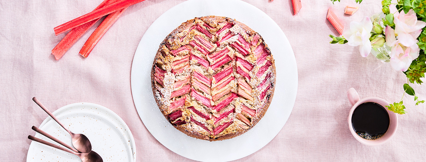 Manteli-raparperikakku valkoisella tarjoilulautasella. Kakku on koristeltu kalanruotokuvioihin asetetuilla raparperipaloilla. Kakun päälle on lisätty tomusokeria. K-Ruoka kakkuresepti.