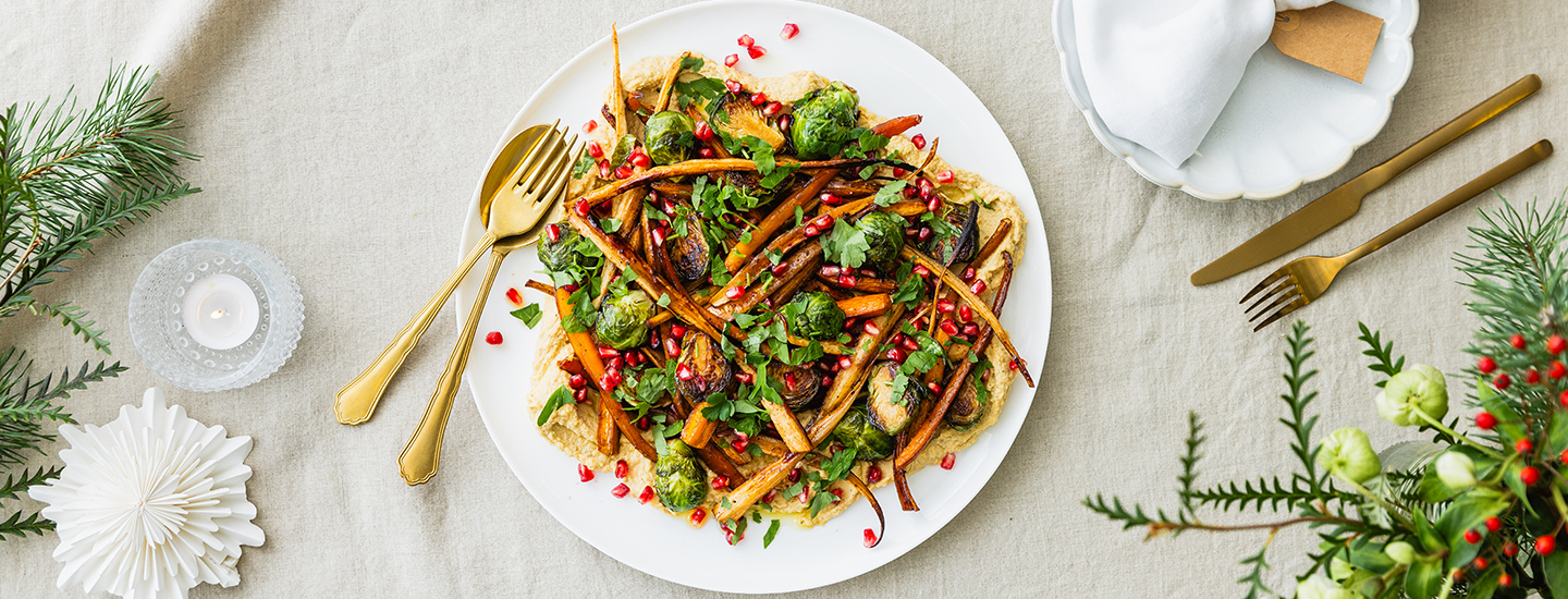 Jouluinen hummusplatter eli hummuslautanen. Isolle pyöreälle lautaselle on aseteltu hummusta, paahdettua porkkanaa, palsternakkaa ja ruusukaalia sekä granaatinomenansiemeniä. Hummuksen vieressä on kultaiset haarukka ja lusikka. K-Ruoka jouluresepti.