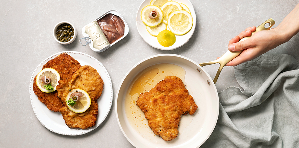 Wieninleike pannulla paistumassa. Leikkeen vieressä on lautanen, jossa on kaksi kypsää wieninleikettä, joiden päällä on sitruunaviipale.