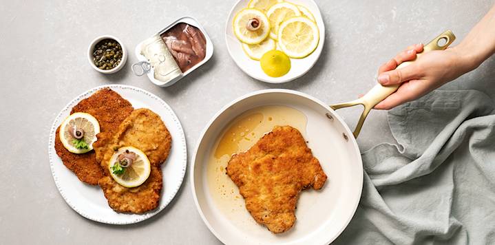 Wieninleike pannulla paistumassa. Leikkeen vieressä on lautanen, jossa on kaksi kypsää wieninleikettä, joiden päällä on sitruunaviipale.