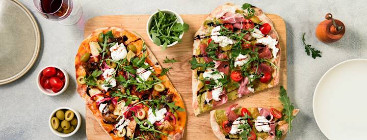 Kaksi pinsa-pohjaista pizzaa puisen leikkuulaudan päällä. Pinsa on ilmava, focacciamainen pizzapohja. Yhden pinsa-pohjan päälle on lisätty parmankinkkua, burrata-juustoa ja kasviksia. Toisen pinsa-pohjan päälle on lisätty erilaisia antipastoja ja burrata-juustoa. K-Ruoka pizzaresepti.