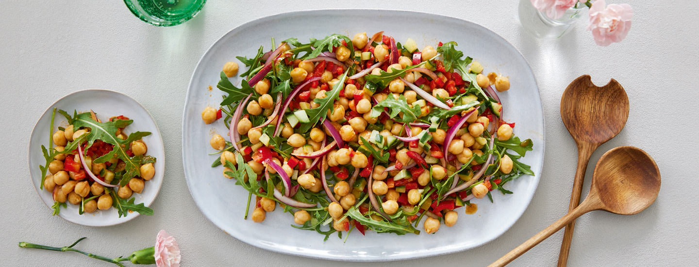 Kikhernesalaatti valkoisella tarjoiluastialla. Vieressä salaattia lautasella sekä ottimet.