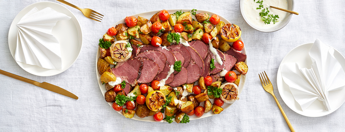 Savulammaspaistia paahdettujen perunoiden ja minikirsikkatomaattien päällä ovaalilla tarjoilulautasella. K-Ruoka pääsiäisresepti.