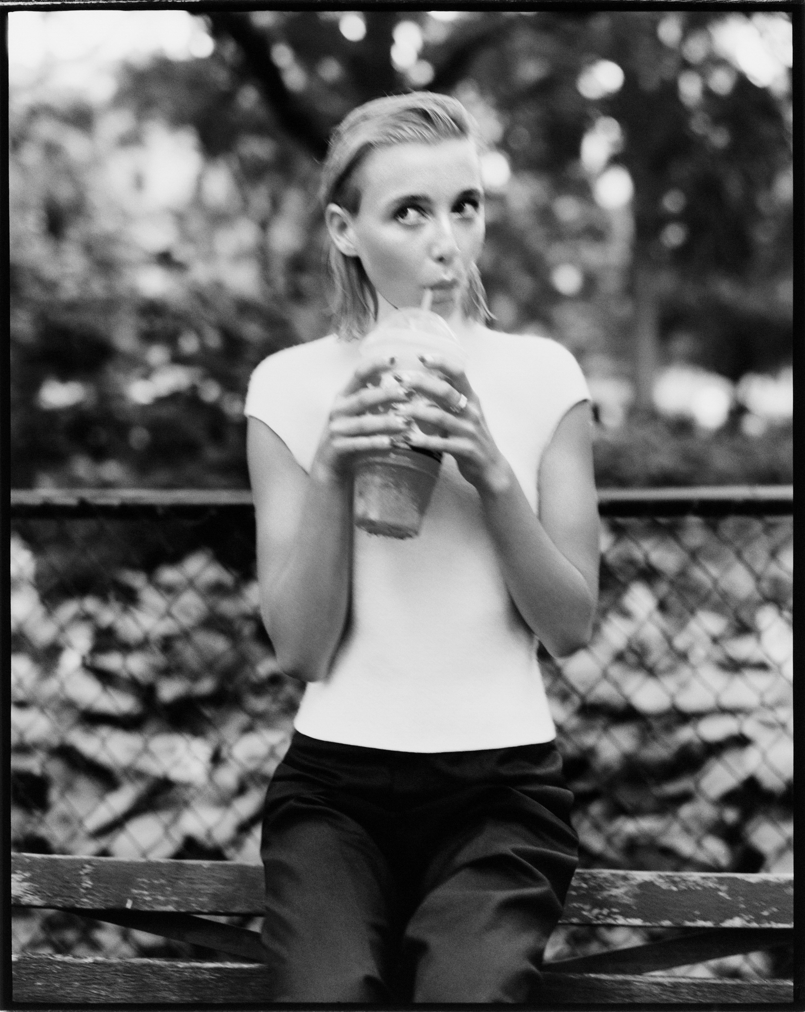 Emma Chamberlain is wearing a simple white t-shirt and black pants. She is drinking what looks to be a milkshake, and is standing in front of a metal fence with trees in the background. 