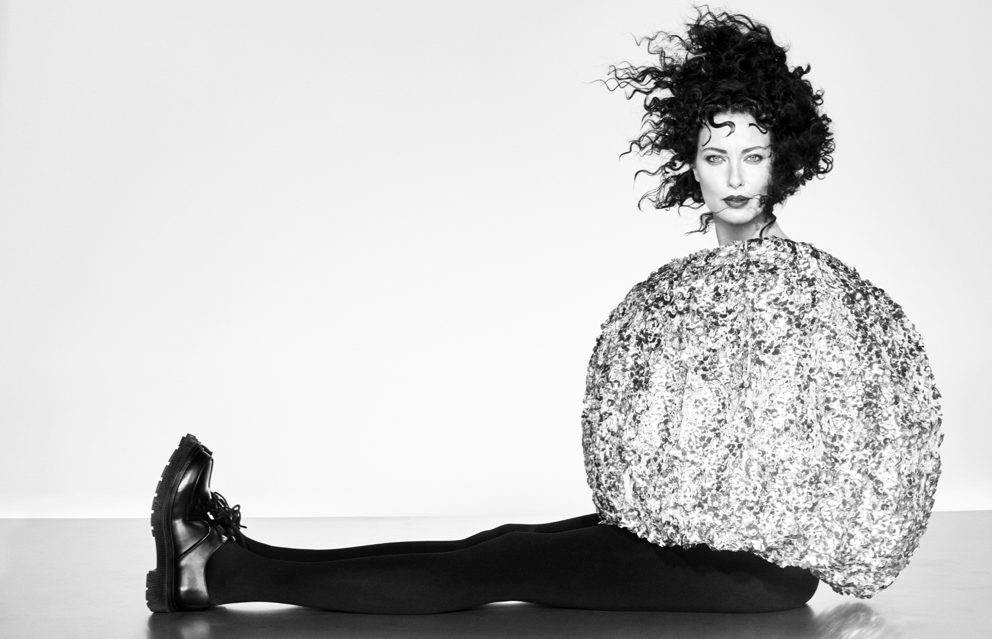 Shalom Harlow is seated on the ground with her legs out in front of her in a straight line. She is wearing a circular shiny garment that contains her hands and arms. She is looking directly at the camera with her curly hair blowing. 