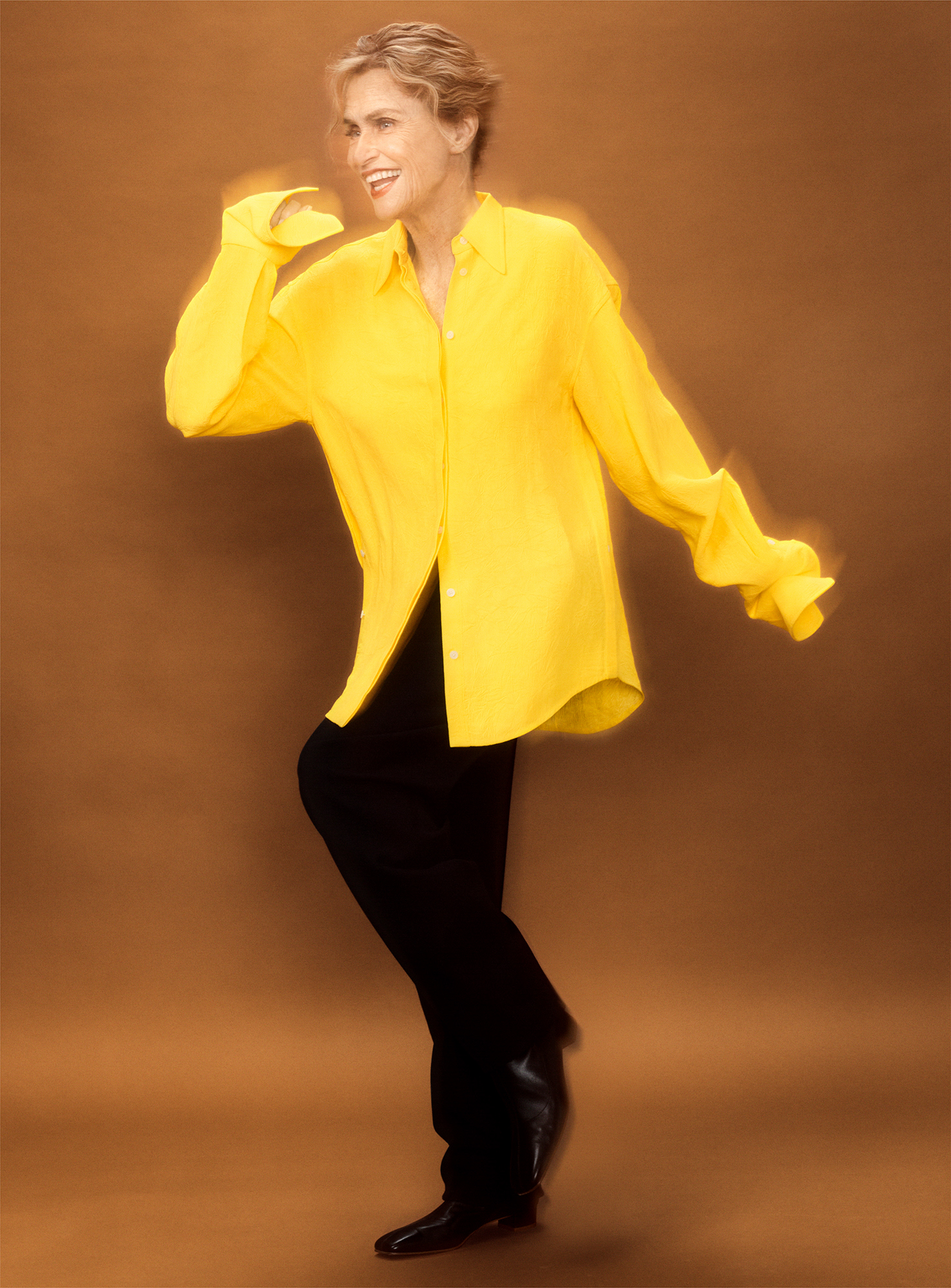 Lauren Hutton has one leg raised and has her arms out as if moving quickly. She is standing in front of a brown background, and is wearing a bright yellow shirt with black pants. 