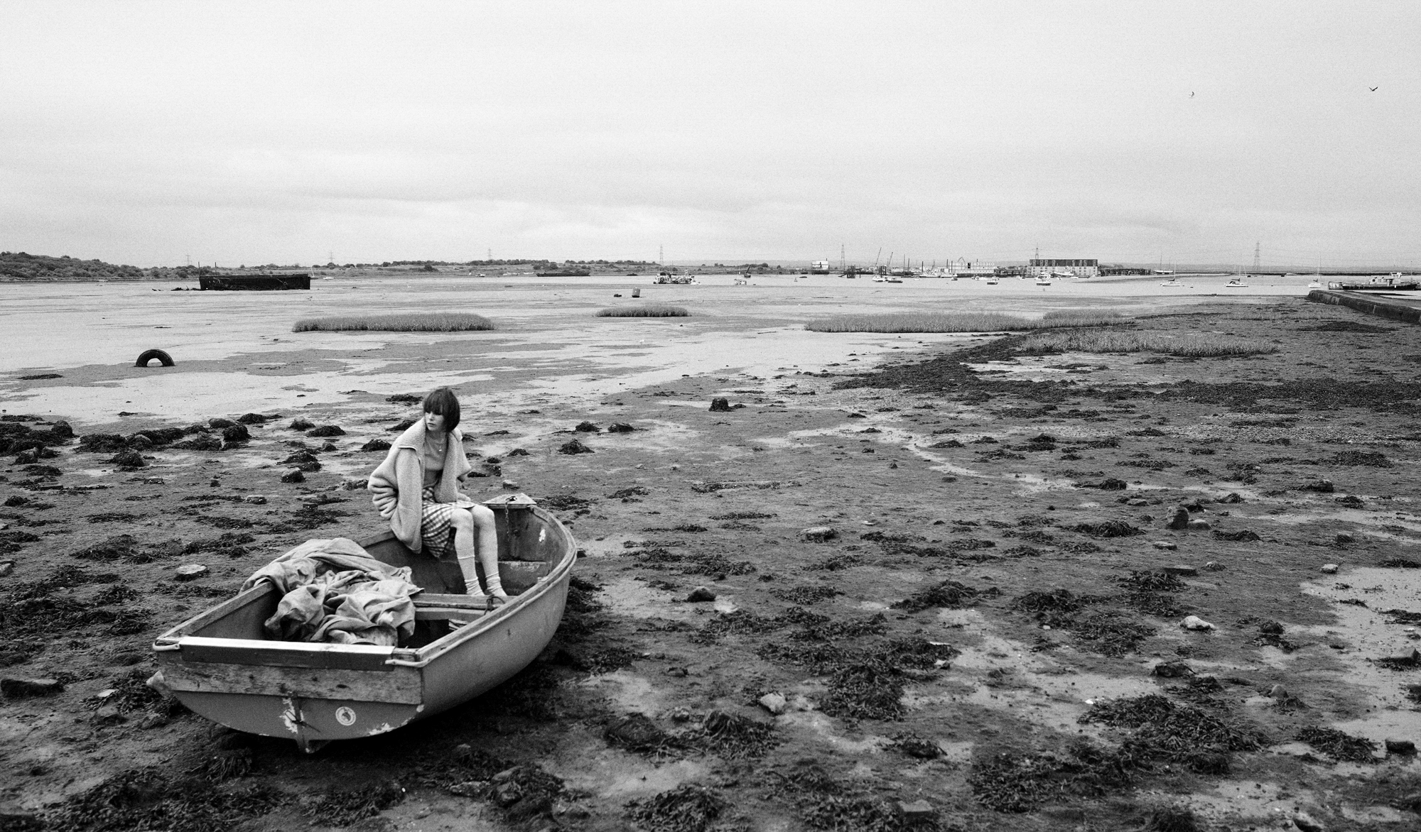 A woman is seated wearing a jacket in a small boat that is on top wet marsh land. The water has receded into the distance, so the boat is on dry land. 