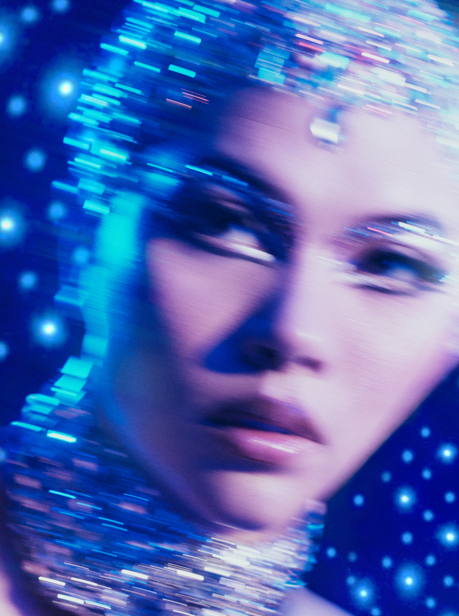 A woman model's face is taking up the majority of the frame. She is wearing a jeweled headpiece and a jeweled necklace. Jewels are featured in the eyeshadow of the look, with graphic eyeliner running along the bottom eyelids. There are blue light orbs in the background, and the entire face is motion blurred. 
