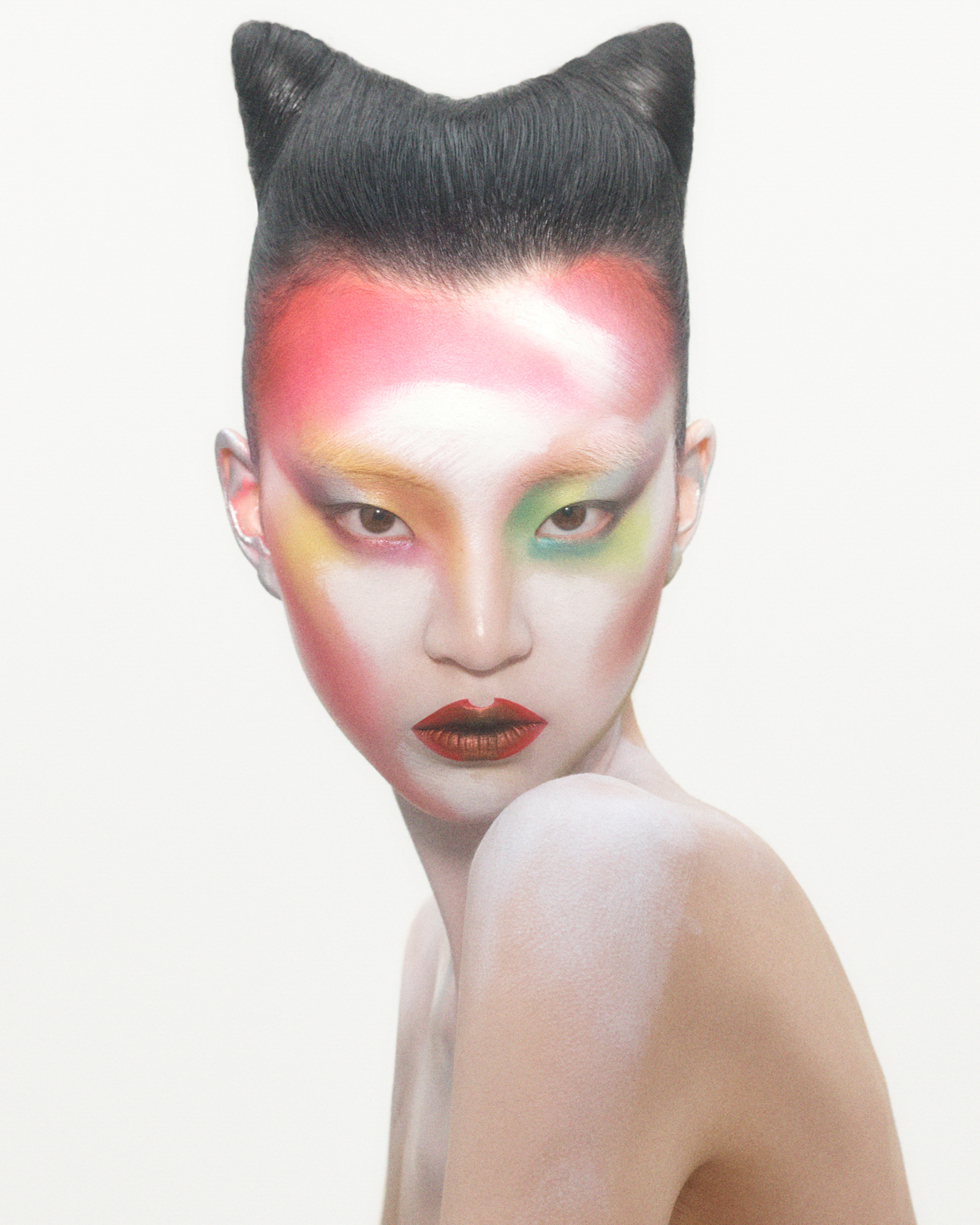 The model is looking straight ahead at the camera. Her face is turned to the side while her shoulders are in profile. Her hair is up in two knots that sit atop her head. She has very colorful makeup on, washes of red frame the face, washes of yellow and green on the eyes, and a bold dark reddish lip. Her face has a white pigment base that extends onto her shoulder. 