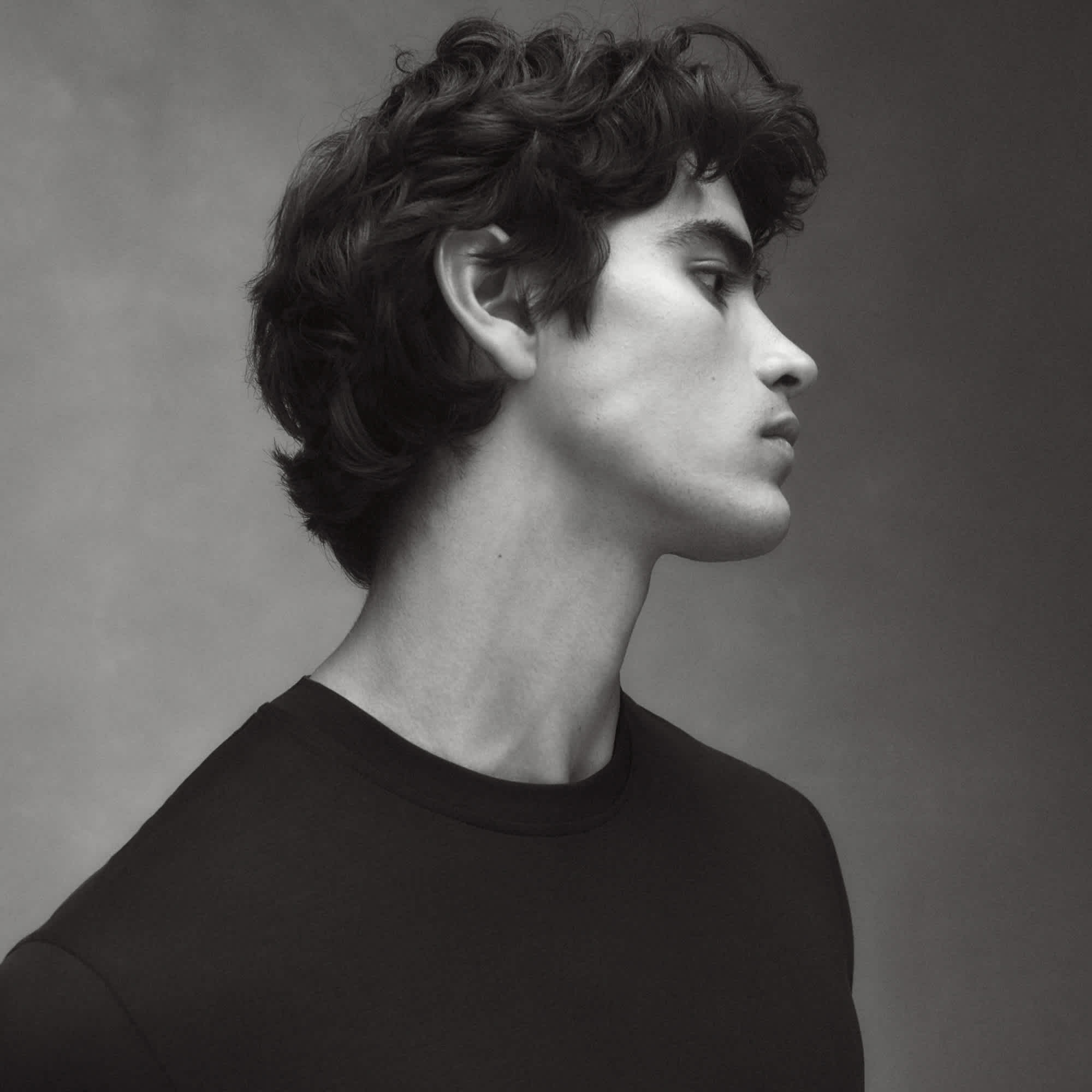 A man with full hair and slight curls is in profile looking off to the right side of the frame. He is wearing a black sweater and is in front of a slightly textured grey background.