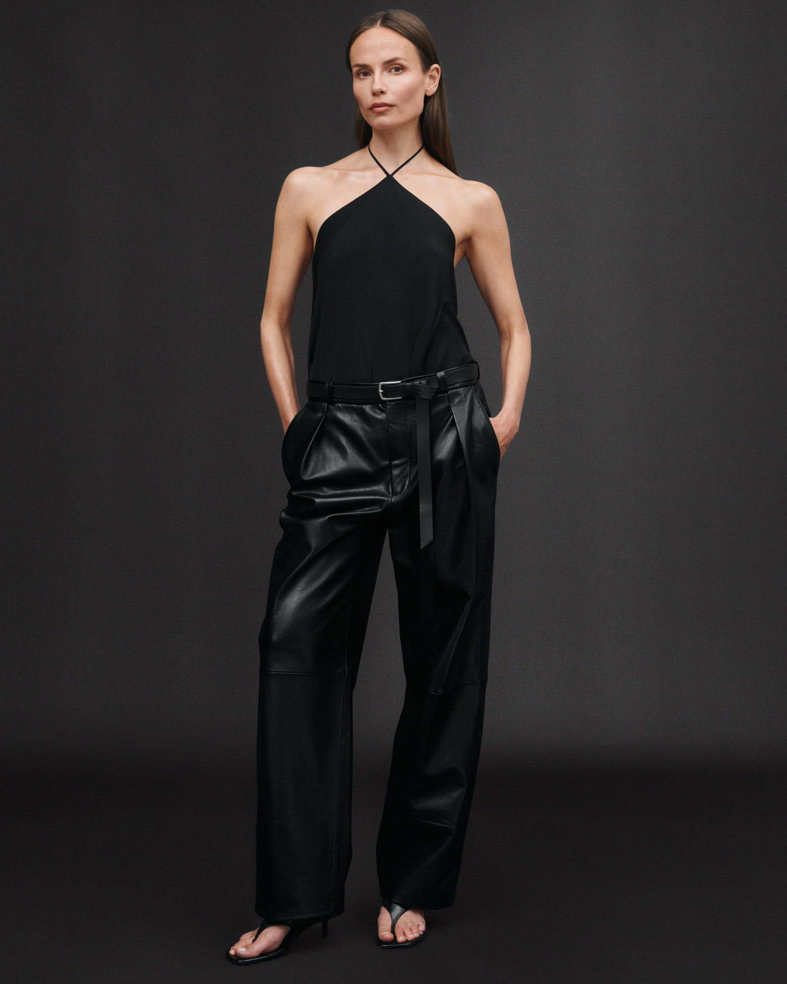A woman model is standing in an all black look against a black backdrop. She is wearing a shoulder less top, and leather pants. Her hands are in her pockets and she is looking directly at the camera. 