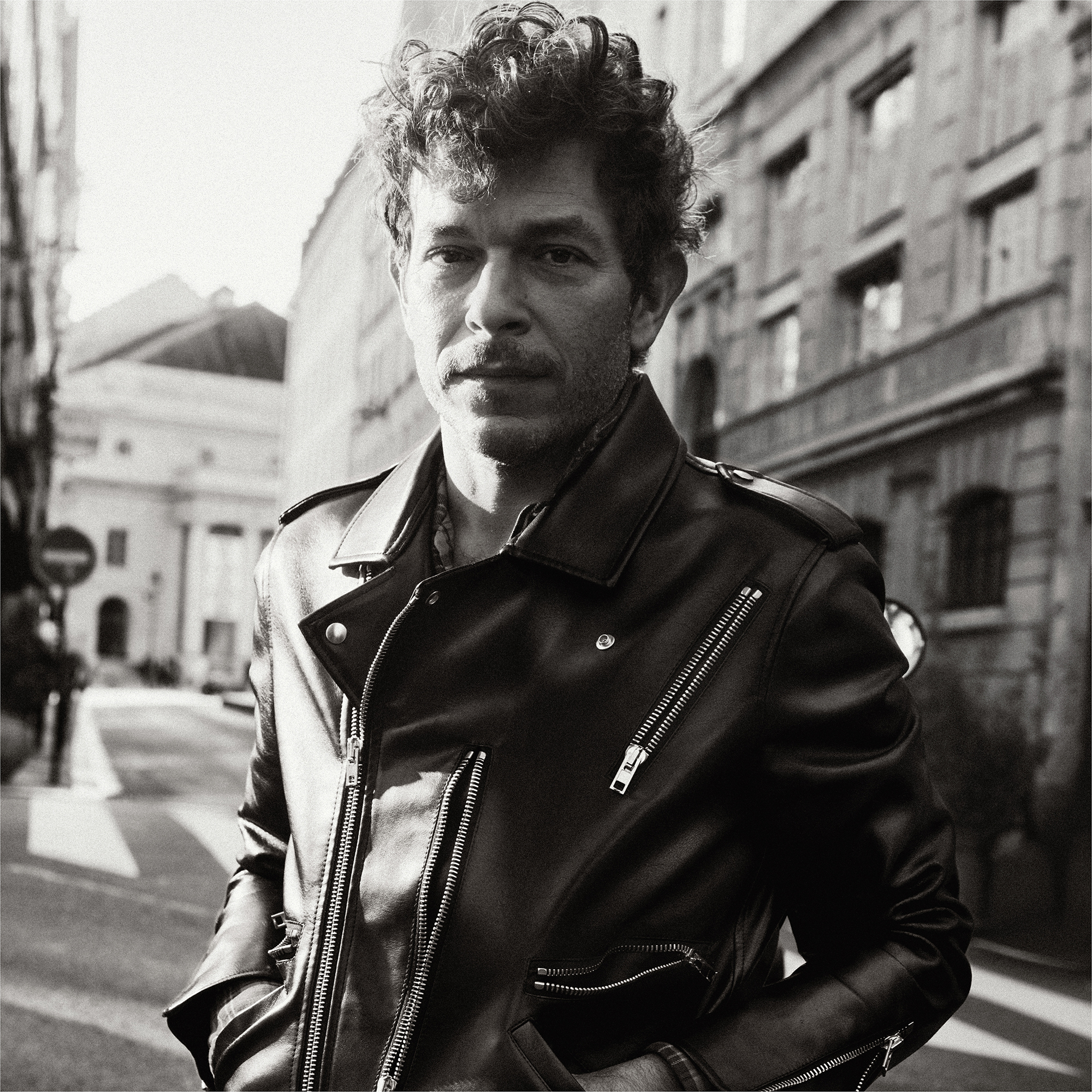 Michael Rider photographed in the streets of Paris. He is wearing a leather jacket with his hair styled up giving volume to his curly hair. He is looking with a straight face at the camera. 