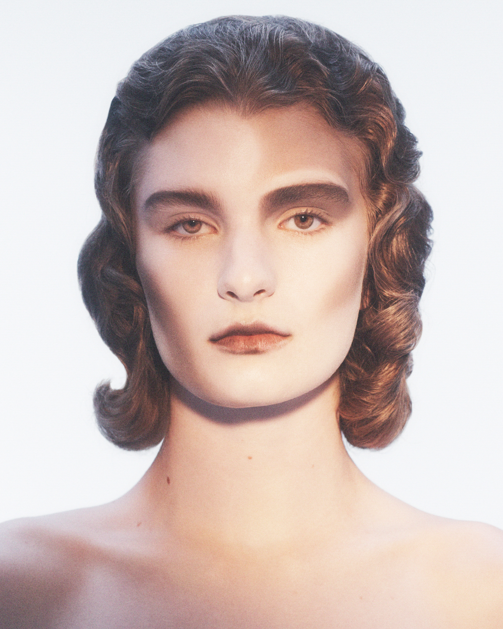 A model is centered in the frame, and is visible from the shoulders up. They have washes of brown sculpting their temples, cheeks, and neck. The brown is carried into the eye sockets and lips. Their hair is in an androgynous sculpted curls that are tight against the face. 