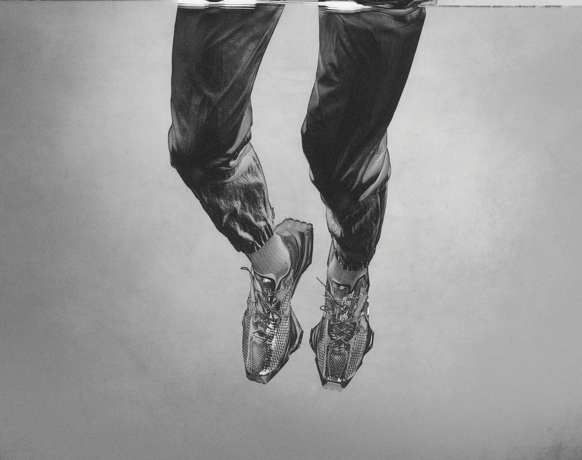 Two legs are emerging from the top of the frame. The legs have an athletic track style pant on. On the feet there are a pair of sporty shoes. A light grey background is behind the legs. 