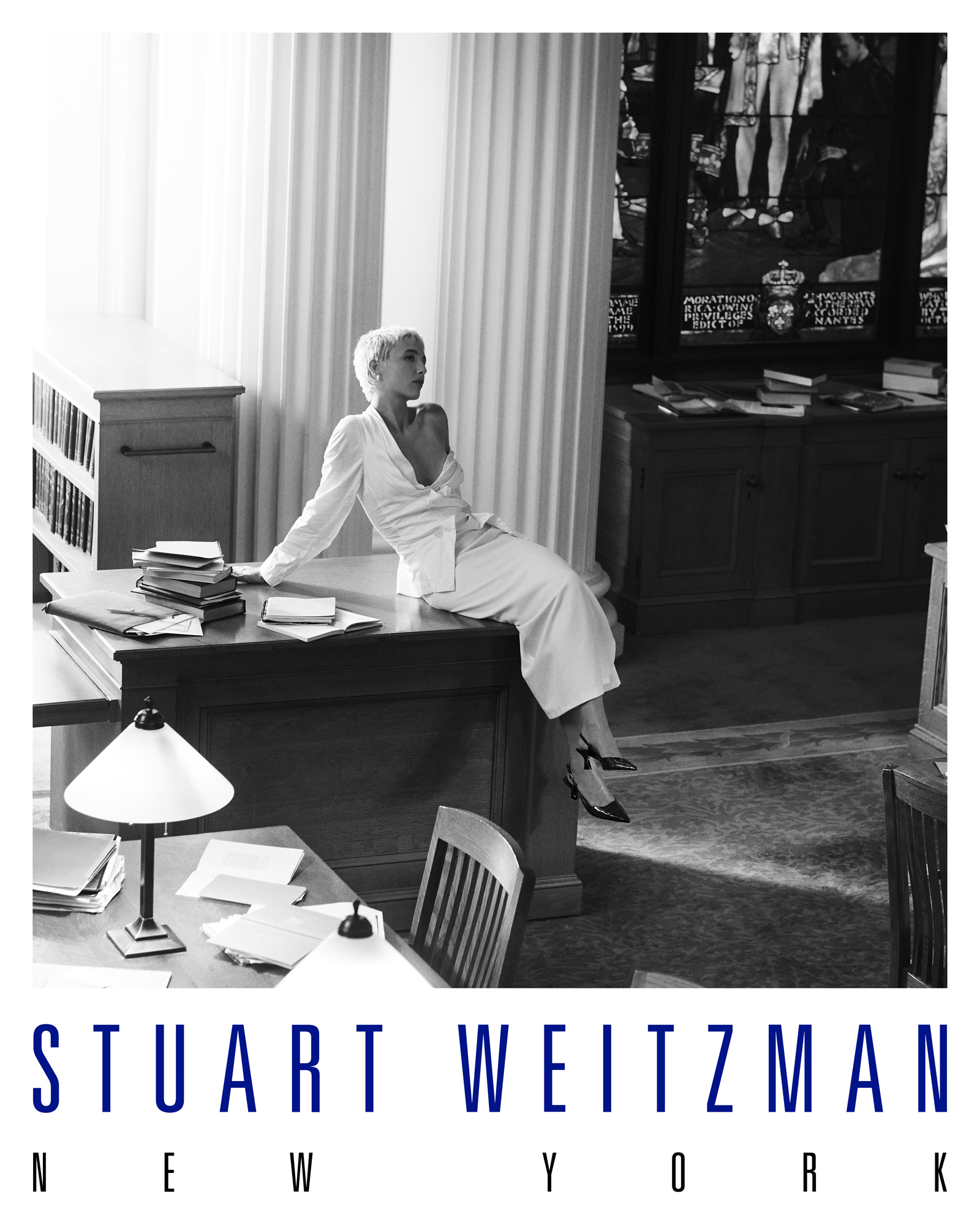 Emma Chamberlain is wearing an all white look with an off the shoulder top and long skirt. She is seated on a wooden desk within a library and is looking off to the right side of the frame. The Stuart Weitzman New York logo is featured at the bottom of the image. 