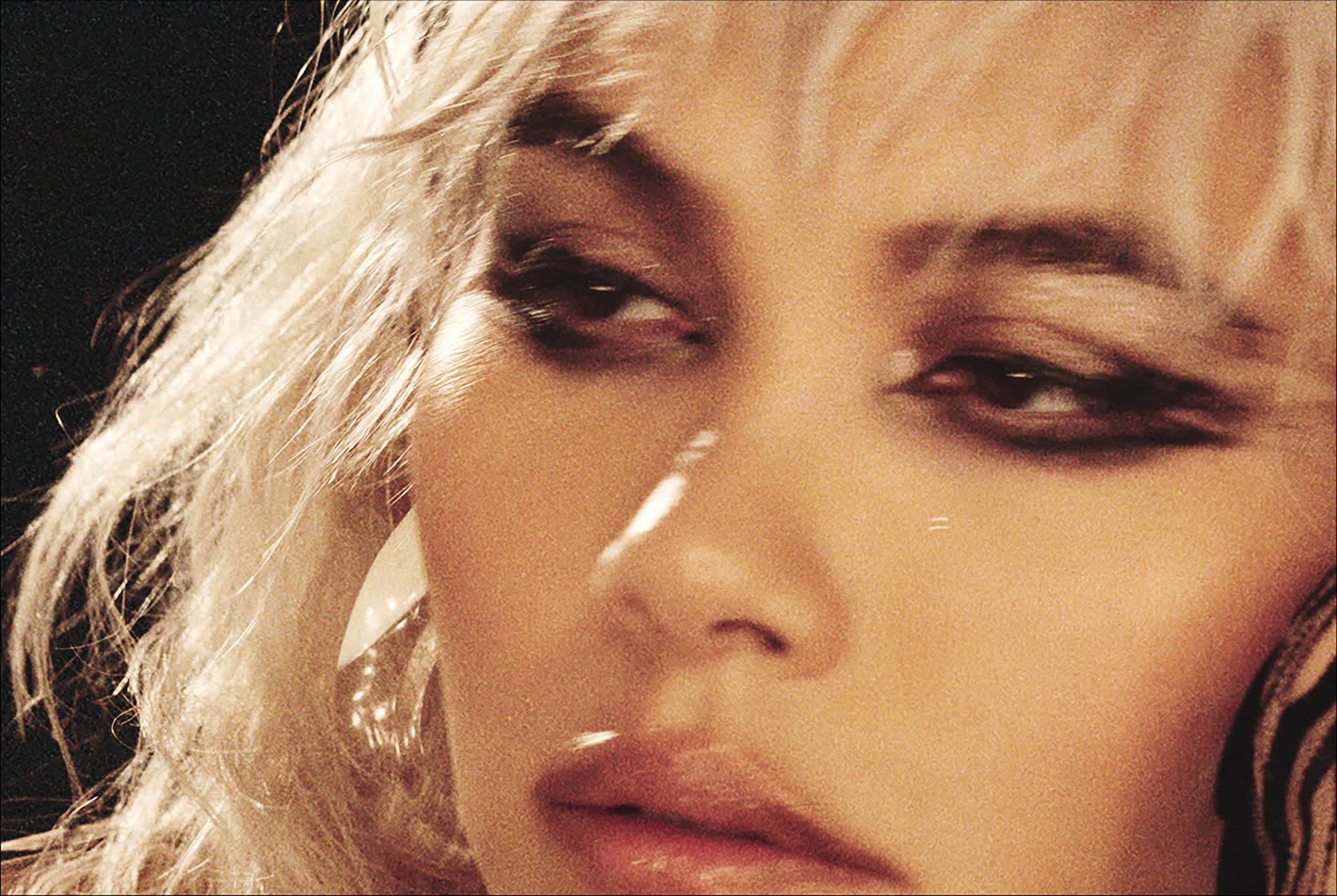 The image is a tight crop of a woman's face. She has blonde choppy hair, and grungy black graphic eyeshadow. Her eyes are looking off to the left, beyond the camera.