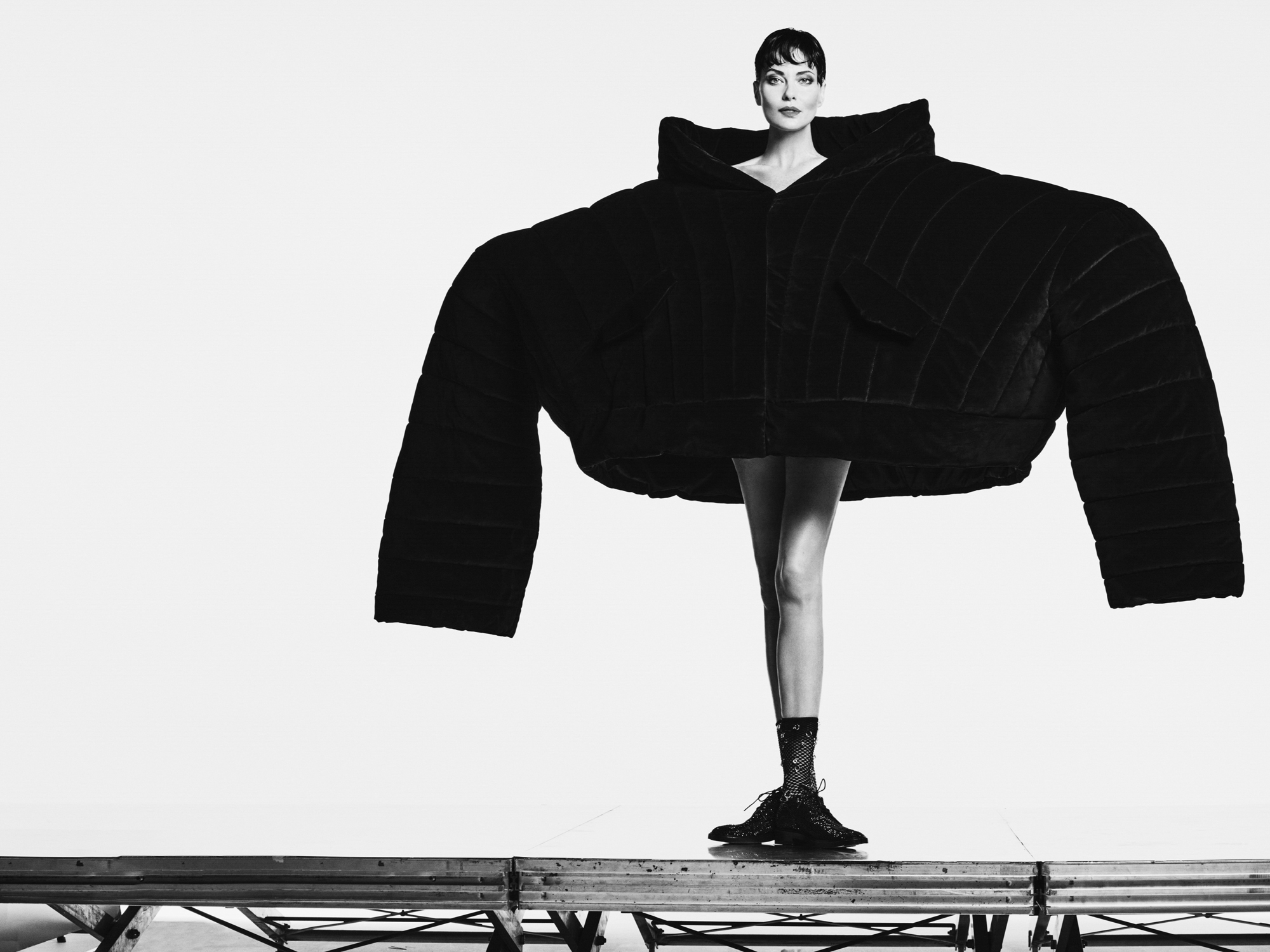 Shalom Harlow is standing on an elevated platform wearing a massive oversized pleated jacket. Her legs are bare and she is looking directly at the camera. 