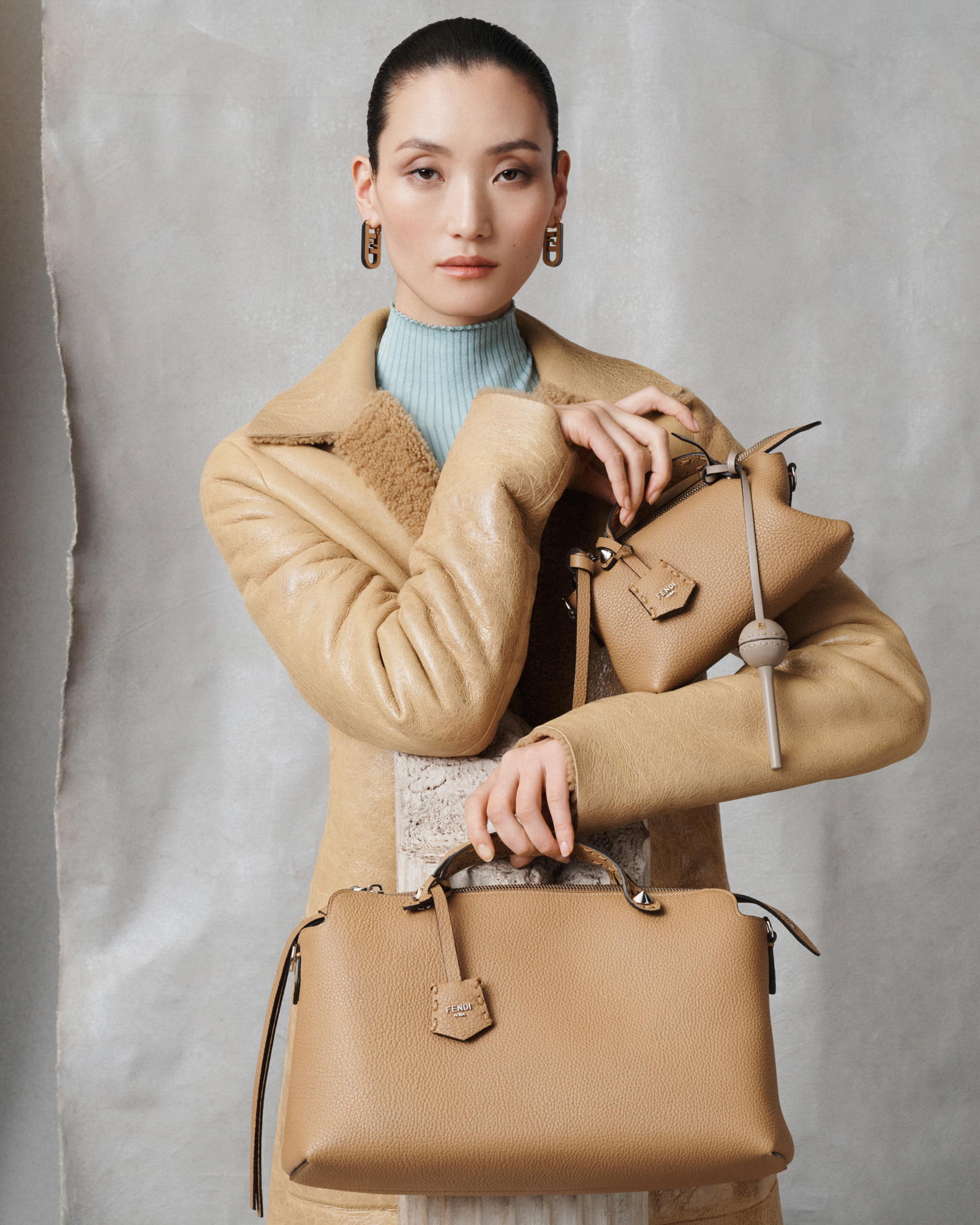 A woman is wearing a light brown leather jacket with a fuzzy lining, and wearing a light blue ribbed top. She is holding two light brown leather Fendi bags, one that is on the large side, and one that is quite small. Her hair is pulled back into a tight bun, and the background is a textured gray paper with the edge showing on the left side of the frame. 