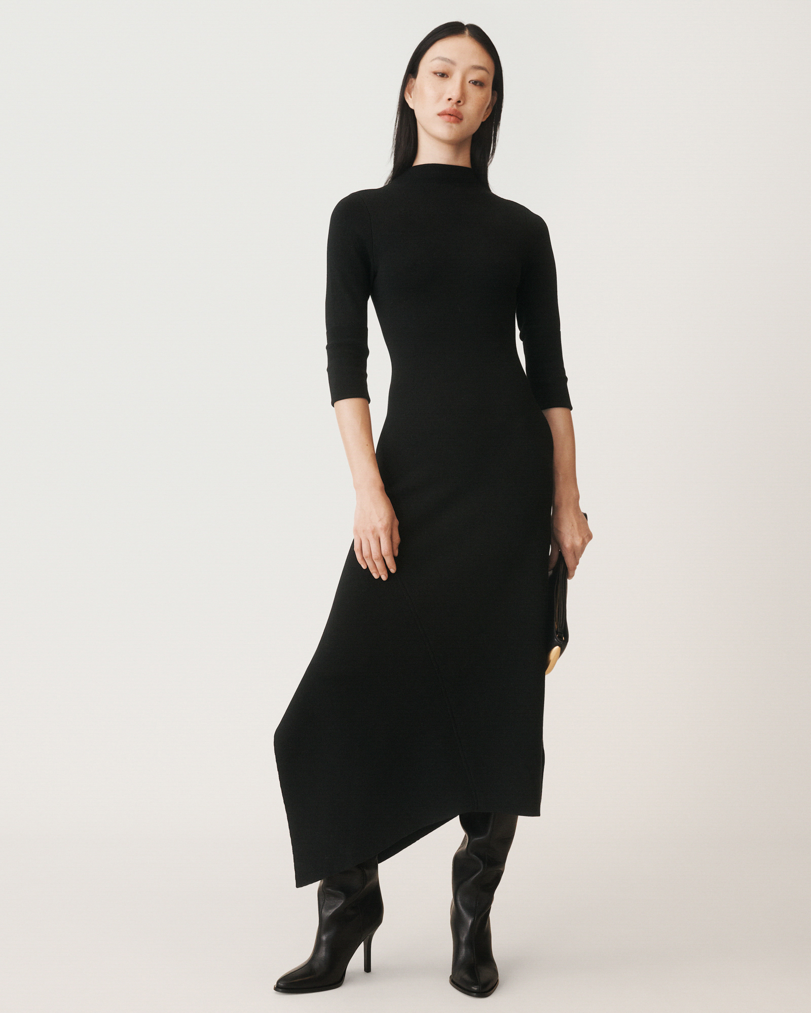 A woman model is standing against a cream white backdrop wearing an all black dress. She is holding a clutch bag and looking towards the camera. 