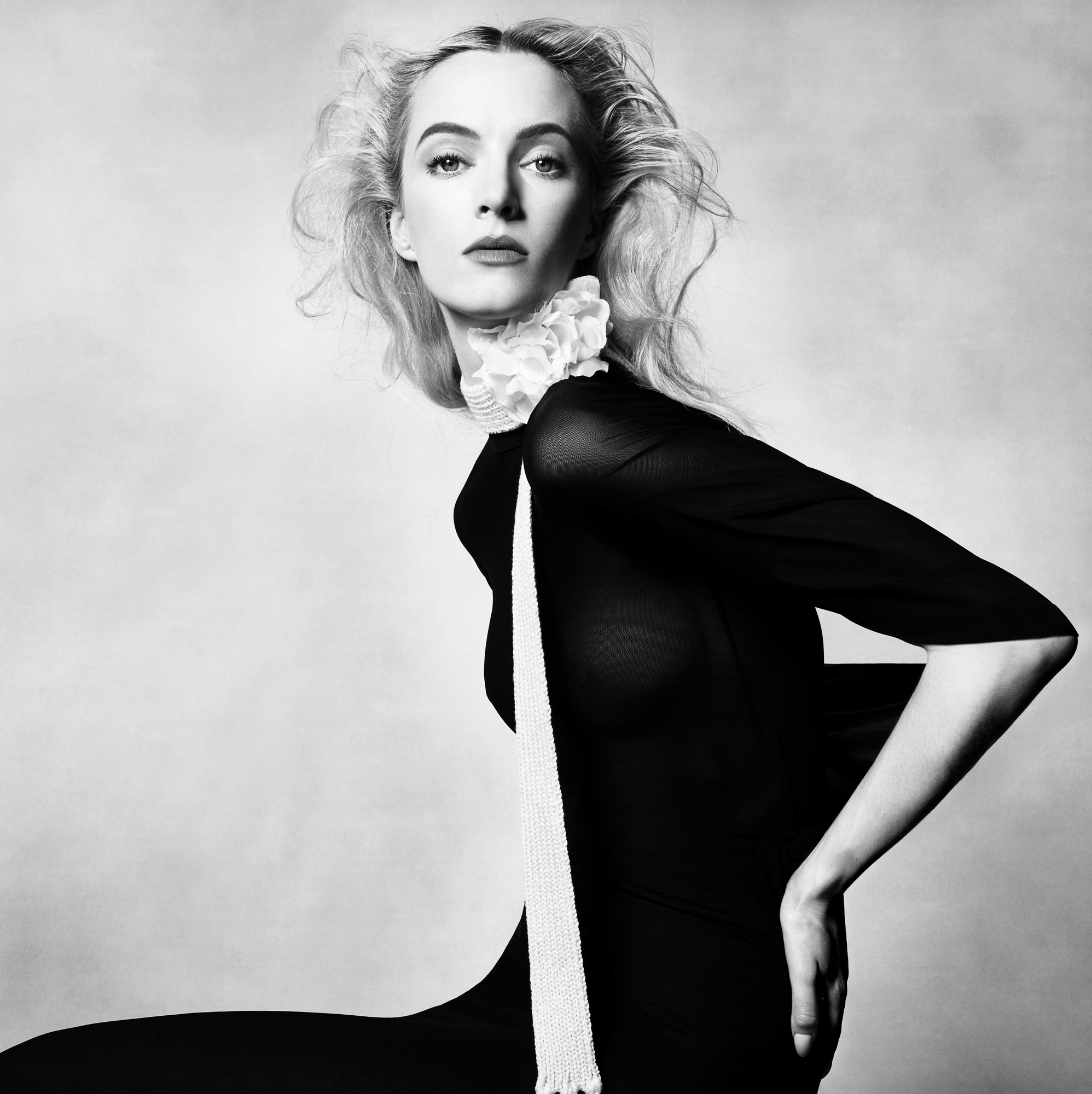 A woman is facing the camera wearing a sleeved black dress. She has a scarf with a floral embellishment at her neck. Her hair is blonde, and being blown away from her face. 