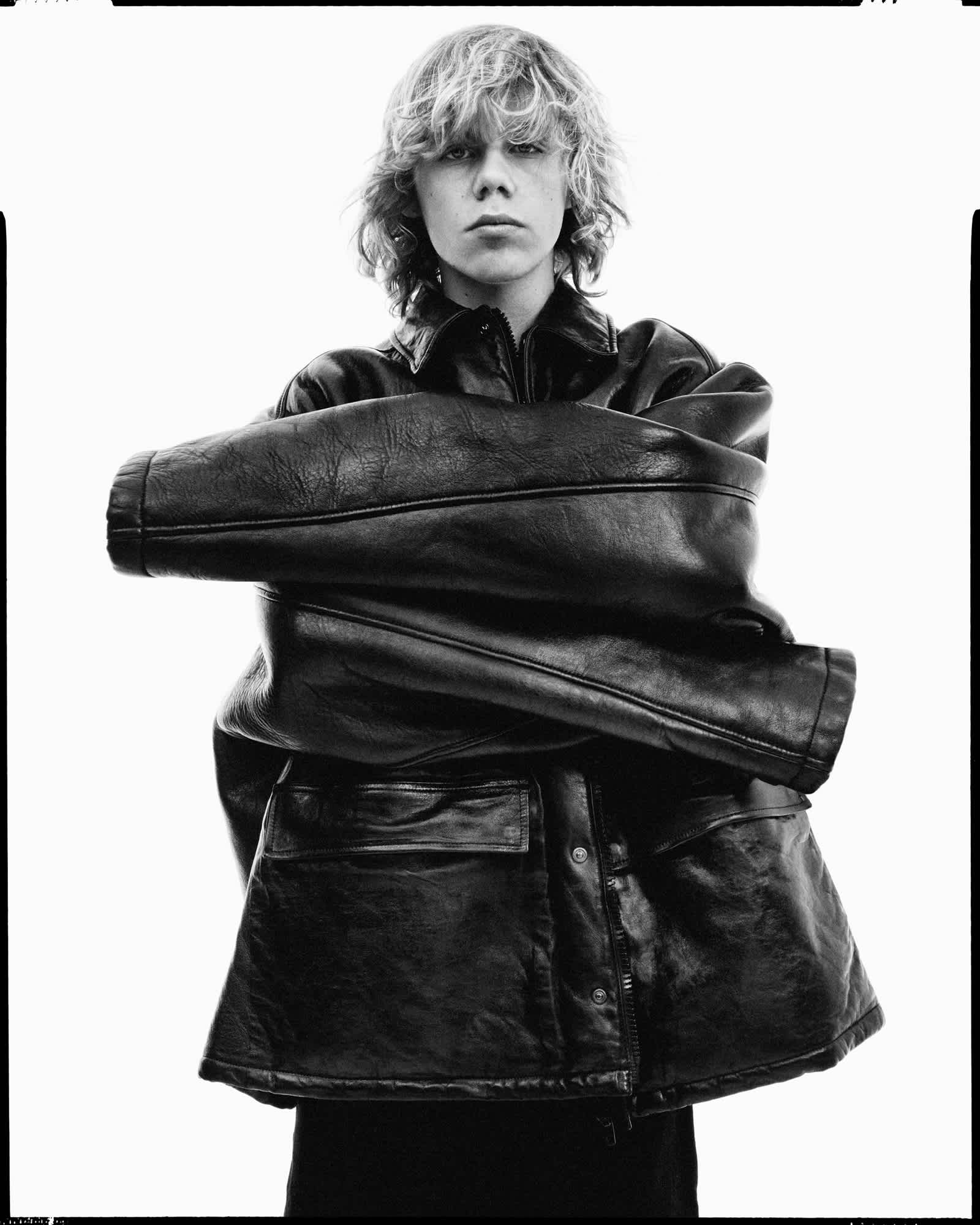 Kid Laroi is standing in the center of the frame wearing a large oversized leather jacket. He is crossing the sleeves in the center of his torso. The entire background is illuminated with a pure white light.