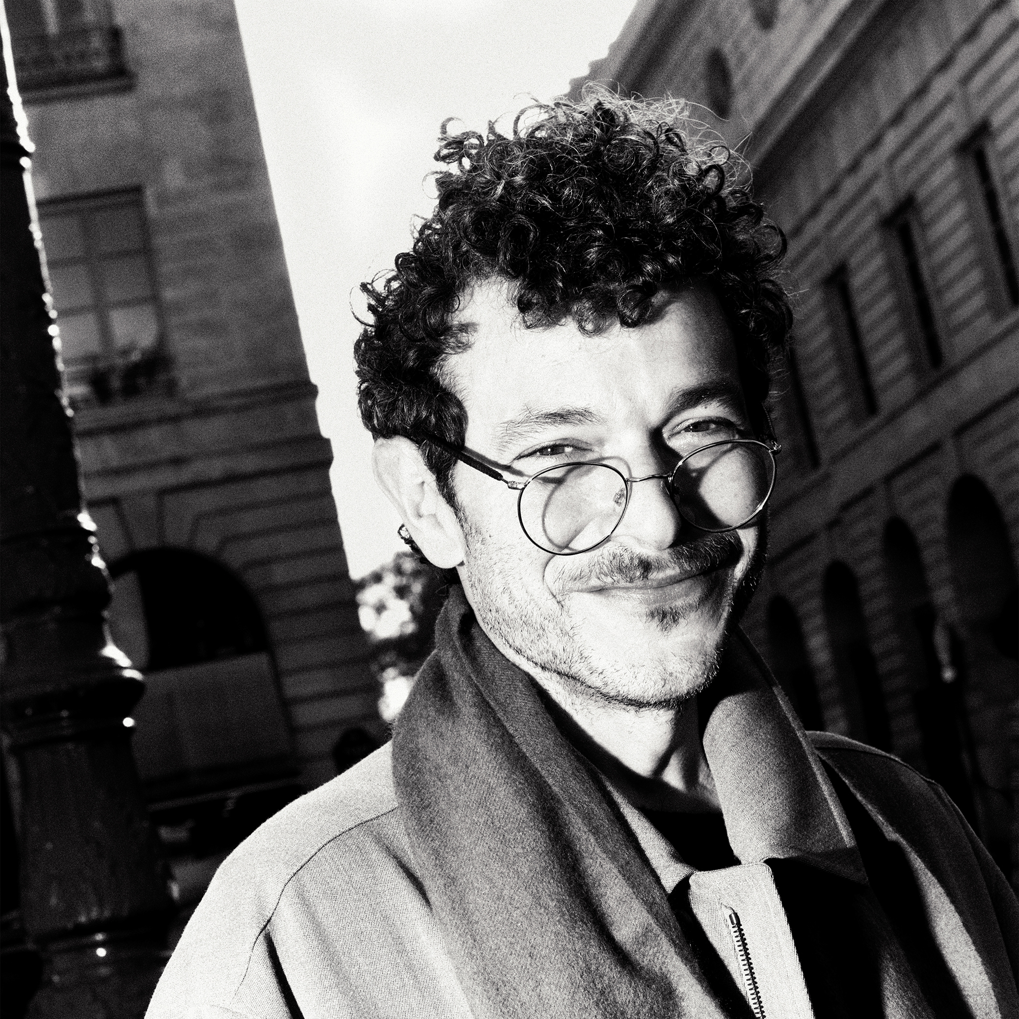 Image of Michael Rider shot in the streets of Paris. He is smiling, wearing glasses, trench coat, and scarf. He is looking directly at the camera. 