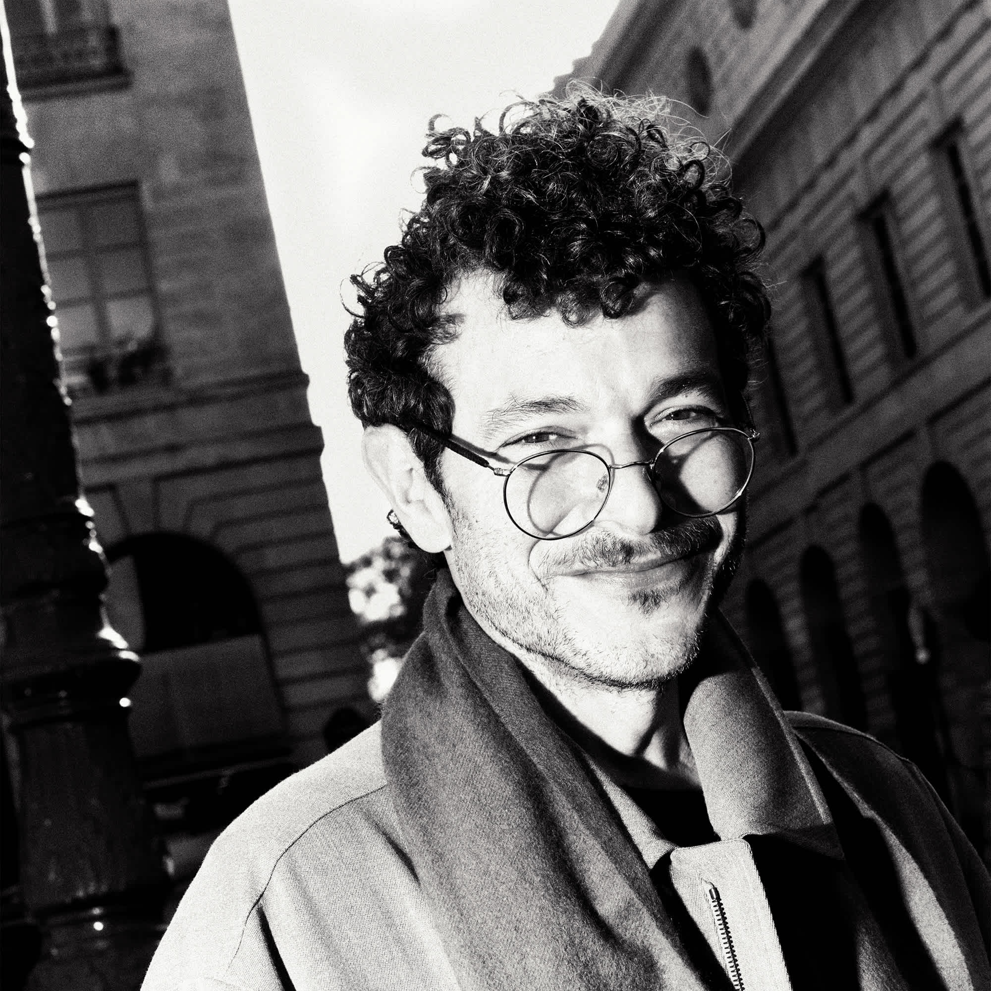 Image of Michael Rider shot in the streets of Paris. He is smiling, wearing glasses, trench coat, and scarf. He is looking directly at the camera.