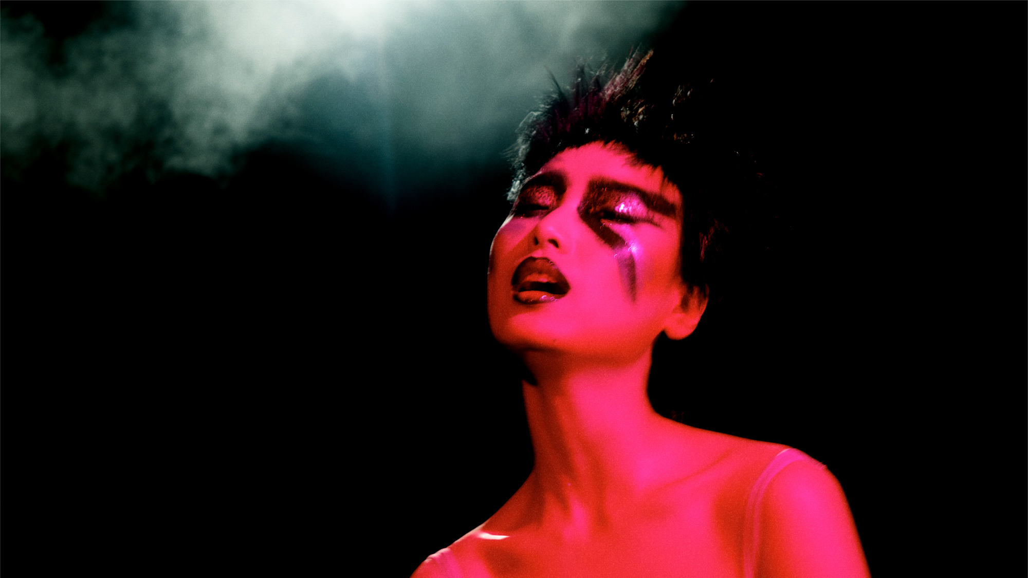 A woman with spiked hair, angular eyeshadow with glitter on the eyelids, and glossy mouth with dark lip liner is being illuminated with pink toned light. A grey smoke cloud is hanging in the area right above the model's head. 