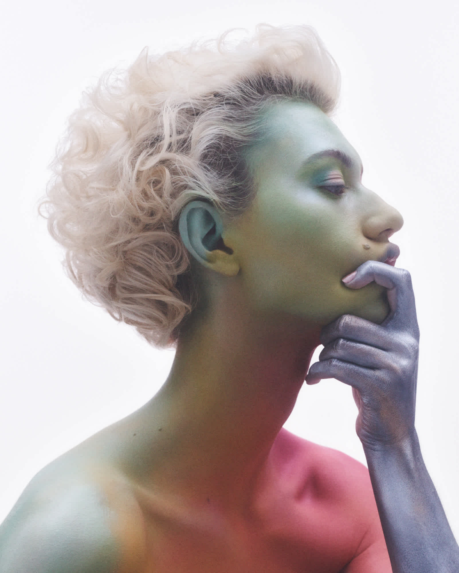 A model is in the frame from the shoulders up, and is turned at a profile. The model's skin has been completely covered by a wash of color, starting with blue in the upper face, yellow and green towards the middle face and neck, shades of red on the shoulder, and blue on the hand and forearm. The model is gripping their chin with one of their hands.