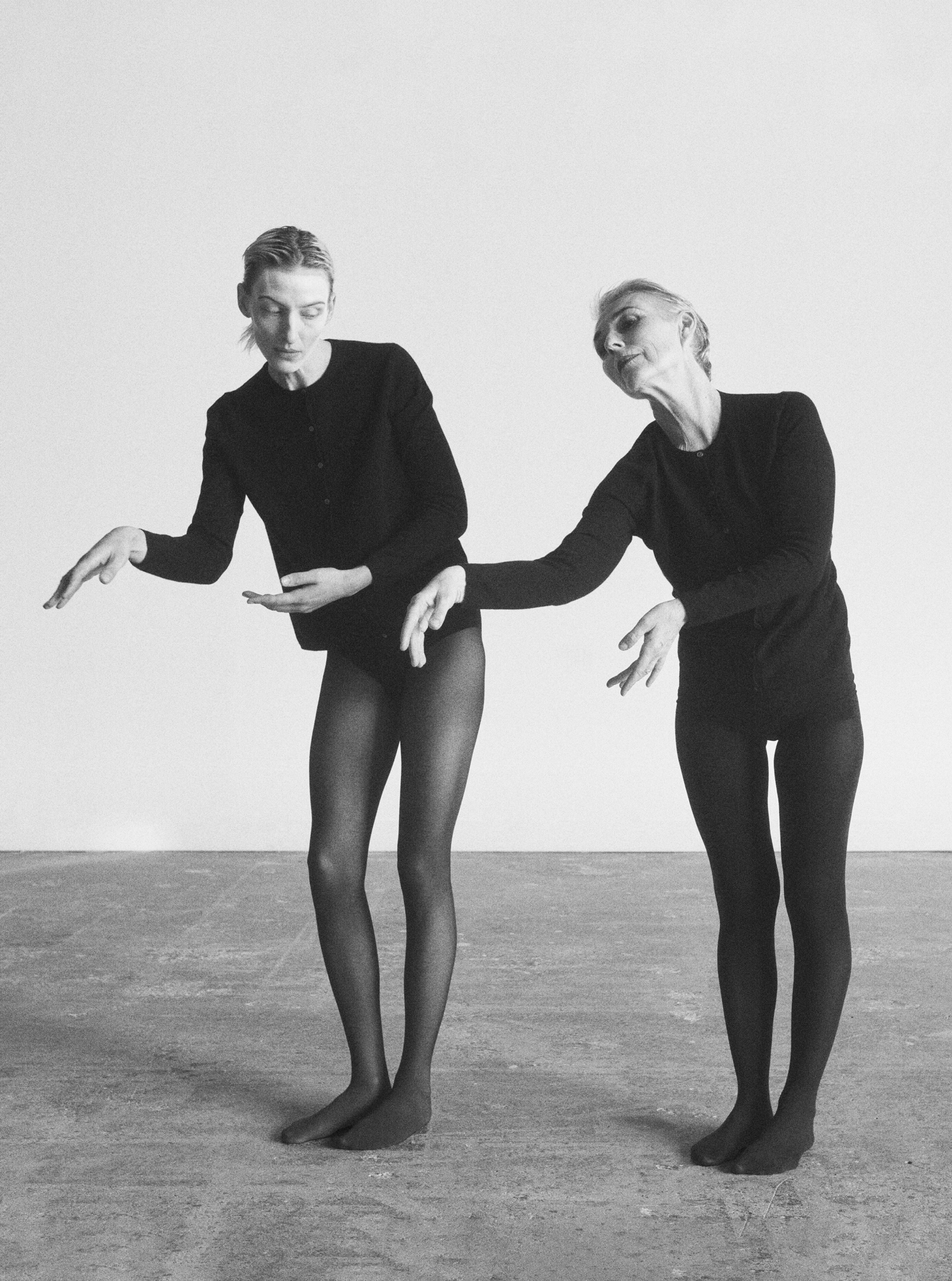 Two models are dressed in all black. They are standing upright and gesturing their hands outward. 