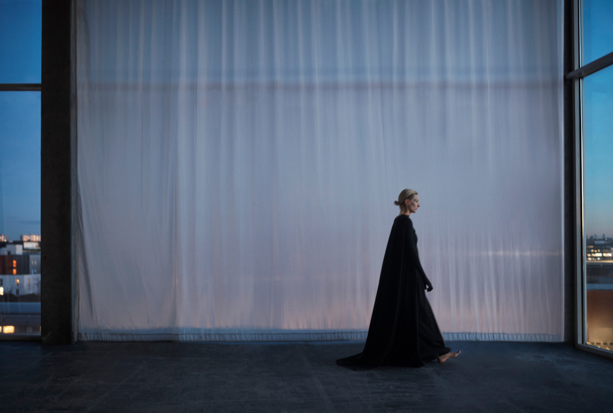 A figure dressed in a full length black dress is walking across the frame towards the right. A sheer curtain is in the background letting in the dusk light. 