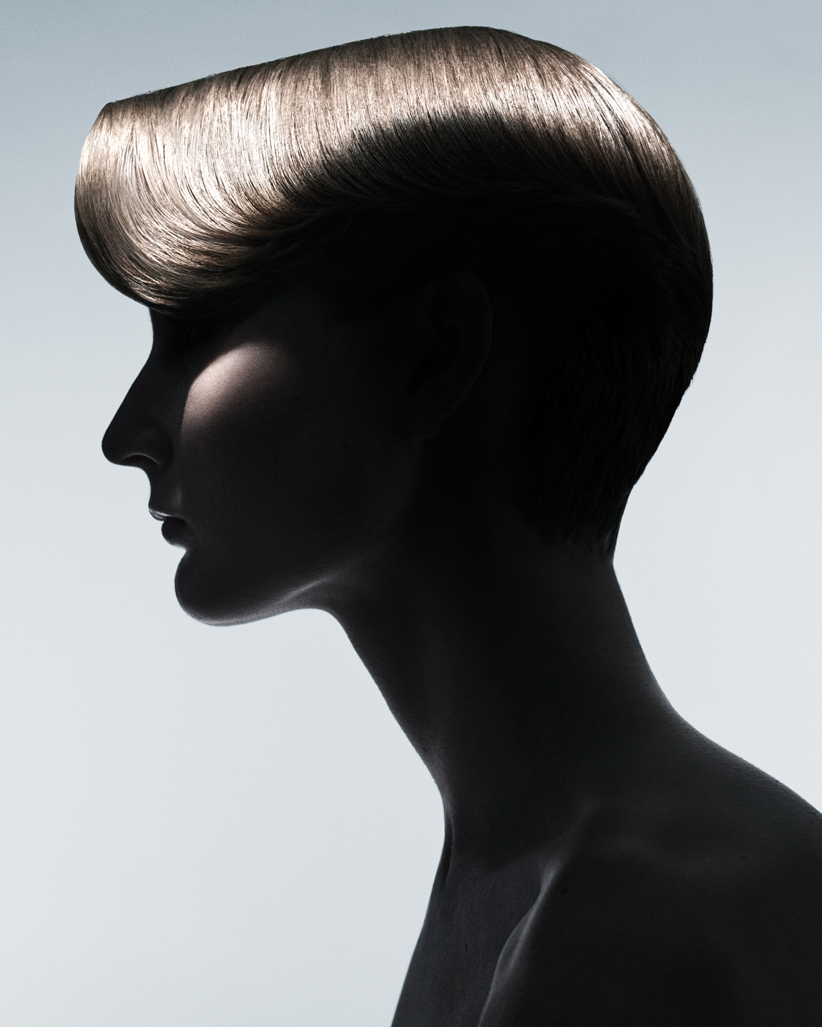 Woman model with short blonde hair is seen in side profile against a light grey / blue background. The model's face is semi obscured by deep shadows. The hair's shine is being illuminated at the top of the head.  