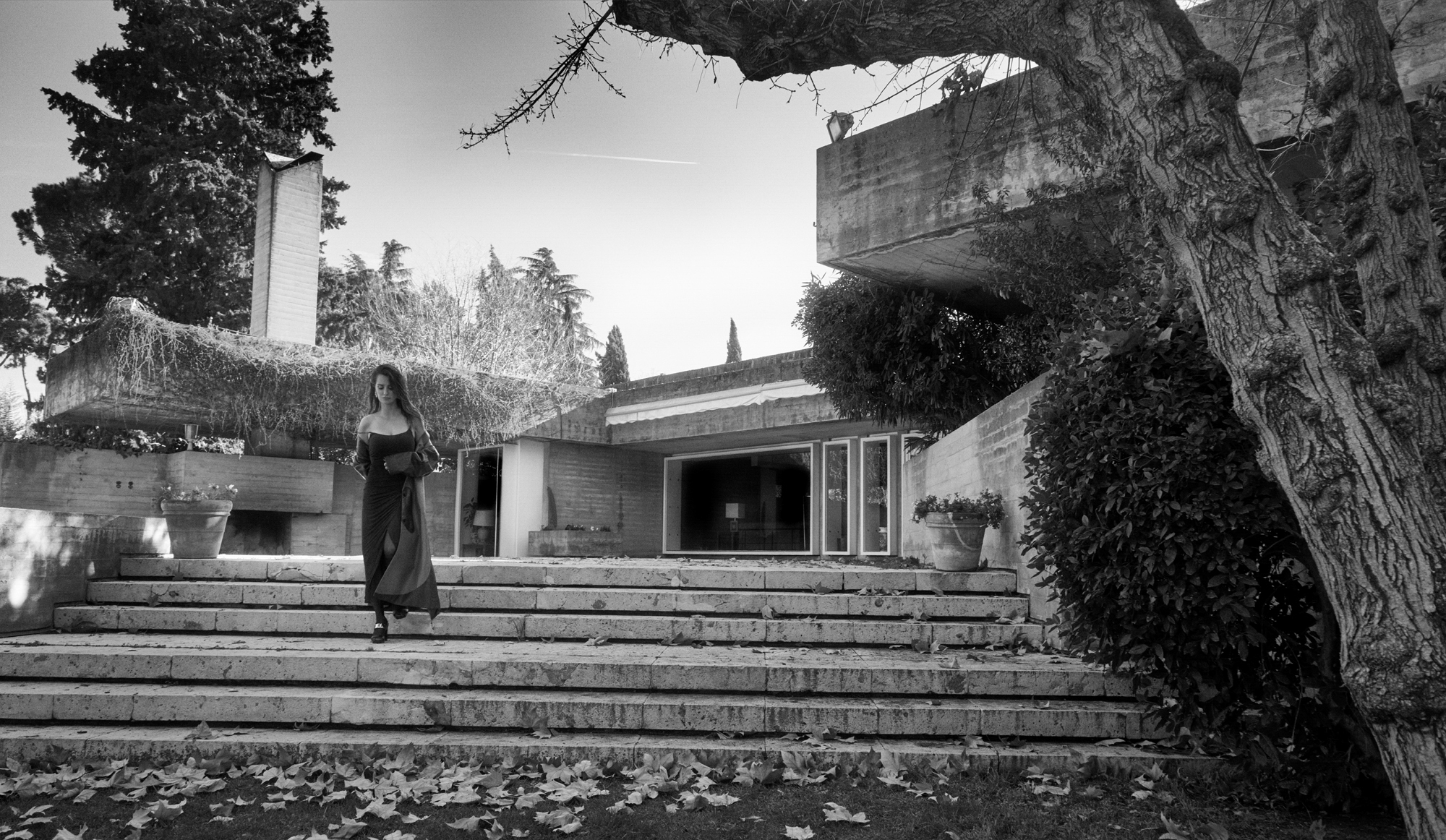Penelope Cruz is walking down the steps to the facade of a Brutalist concrete house. She is wearing a full length dress with a trench coat over the top. 