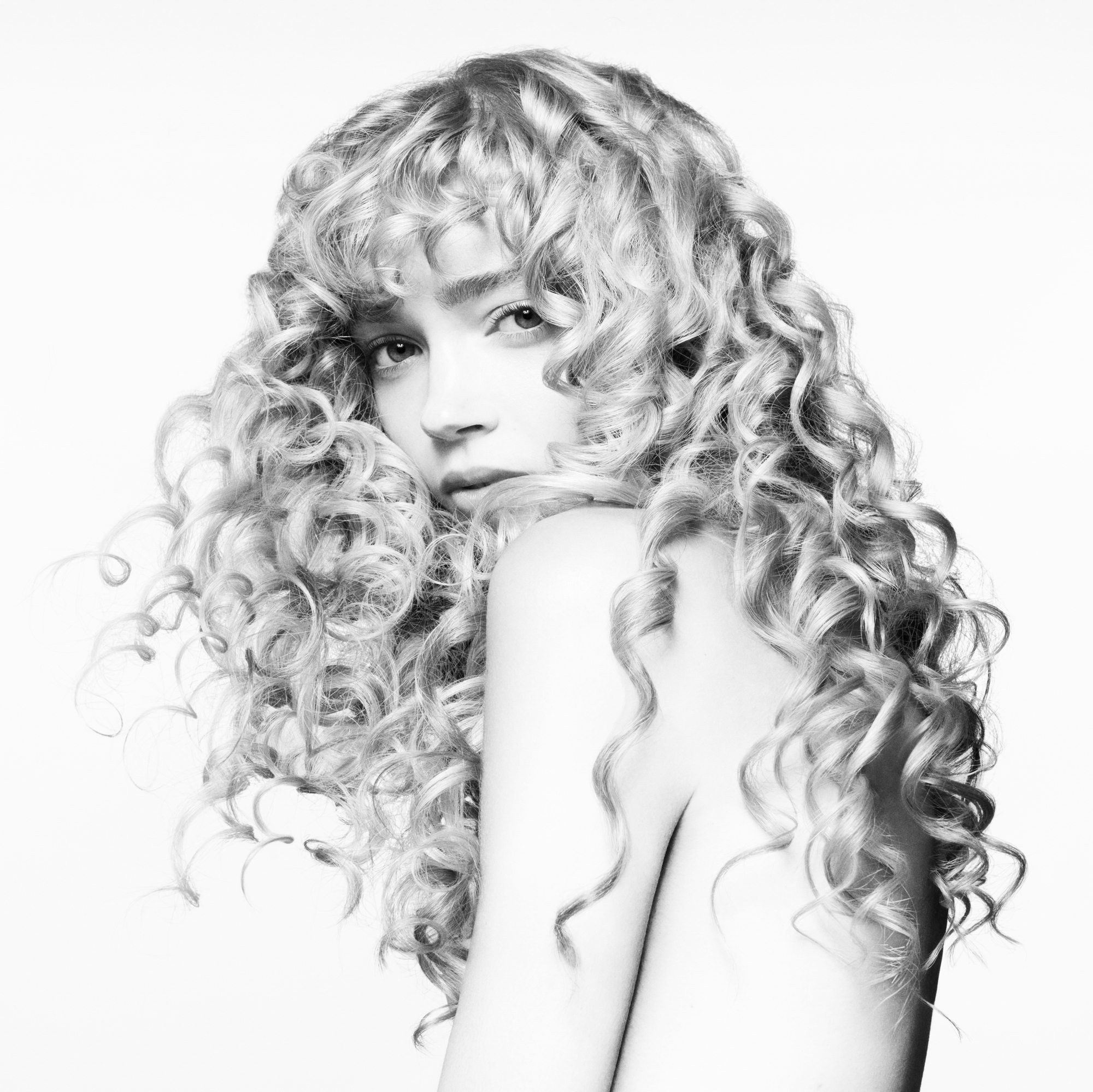Black and white image of a blonde woman with long curly hair. Her face is tucked away behind her shoulder slightly, and she is looking ahead at the camera. Her hair is blowing from behind. 