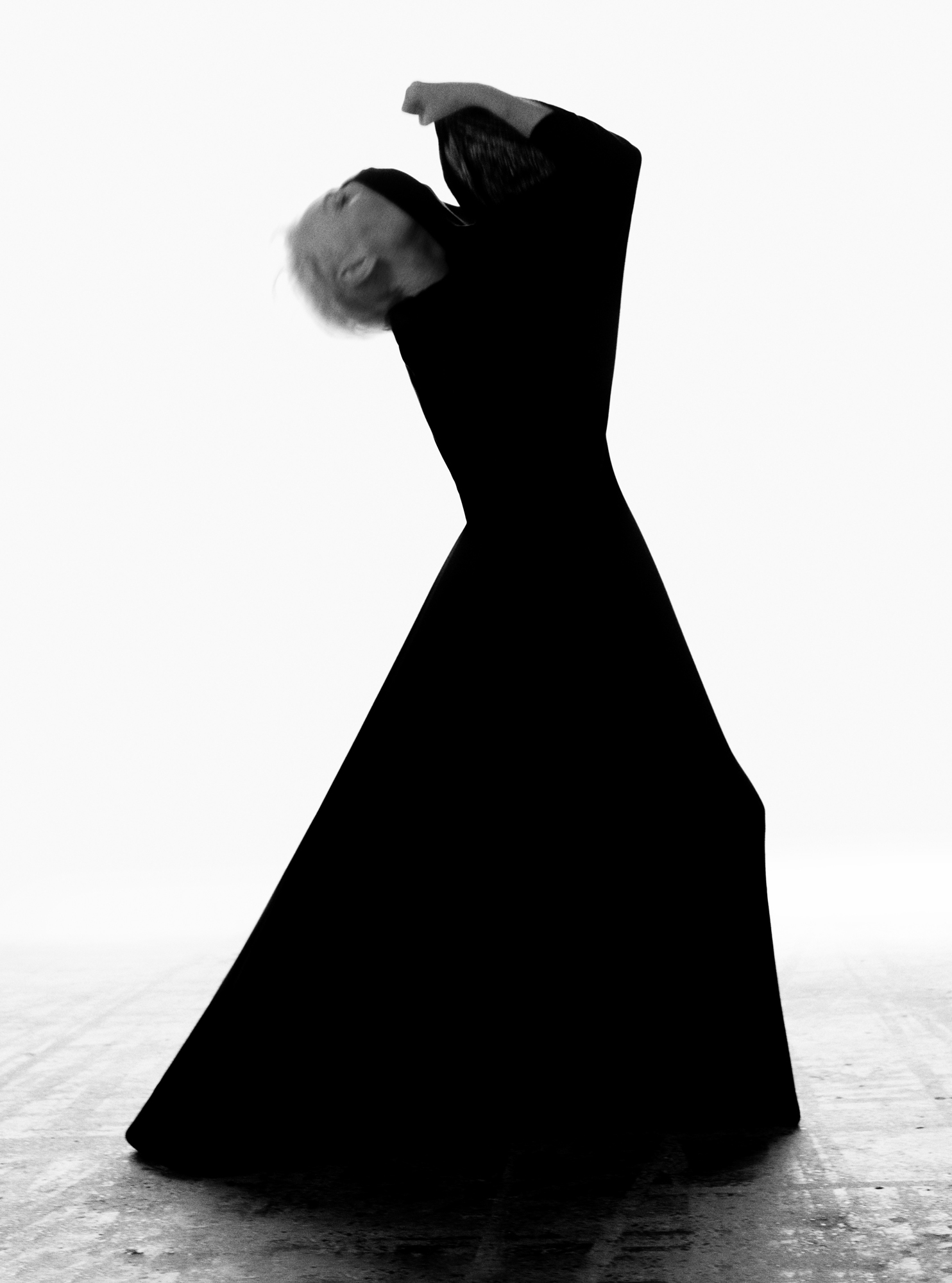 Figure wearing a full length black dress is arching head backwards. Arms are raised slightly above the figure's face. Figure is seen from profile, light is pouring in from the background. 