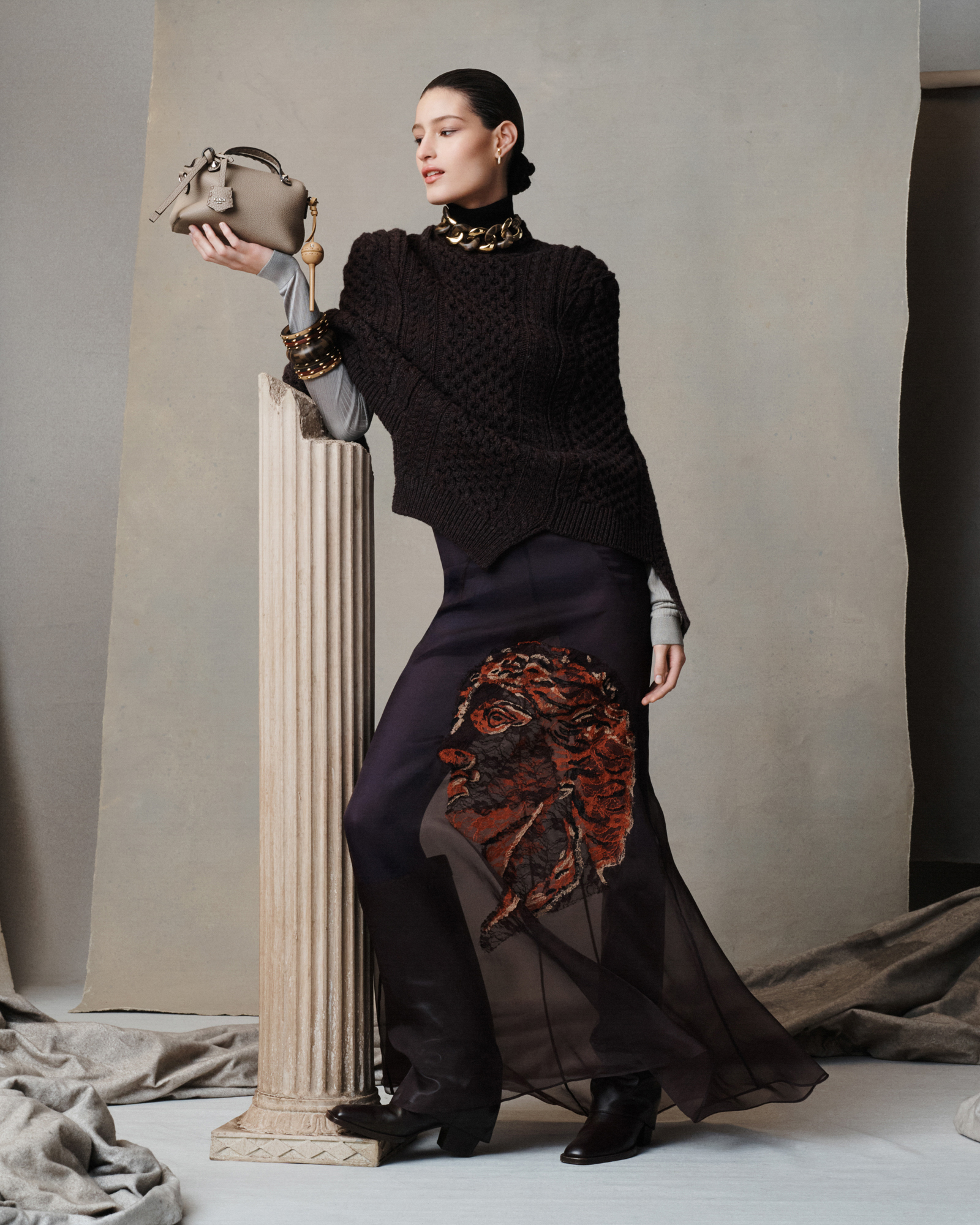A woman is standing in the center of the composition, she is wearing a textured knit sweater, a sheer dress underneath with an embellished Greco-Roman statue head, and black boots. The model is holding up a small brown handbag, her arm resting on a Greek column. On the set there are different tones of grey paper and a grey fabric that is near the model's feet and the Greek column. 