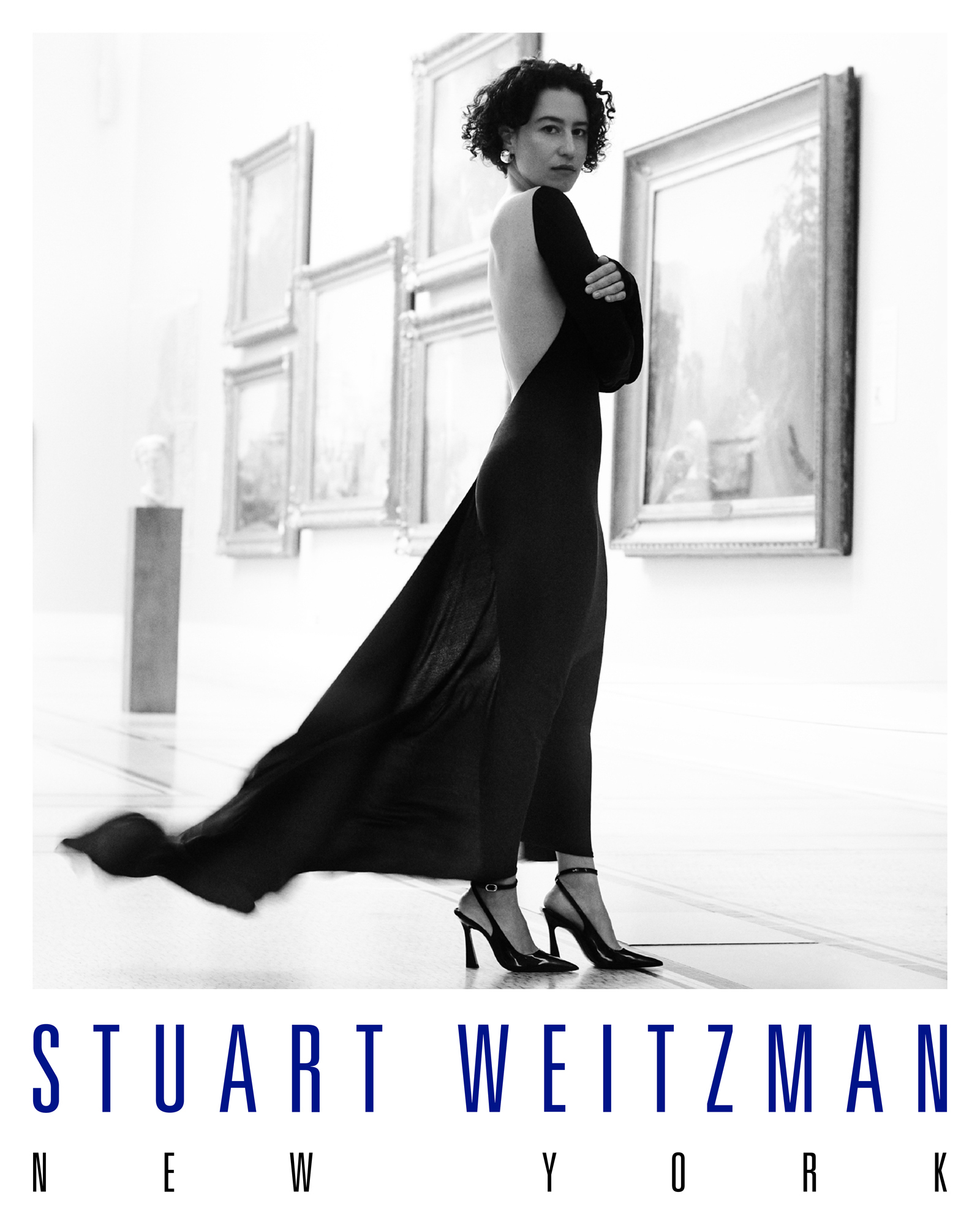 Ilana Glazer is wearing a full length backless black dress with black pumps. She has her arms crossed and is looking towards the camera. She is standing within an art gallery with paintings. The Stuart Weitzman New York logo is featured below the image. 