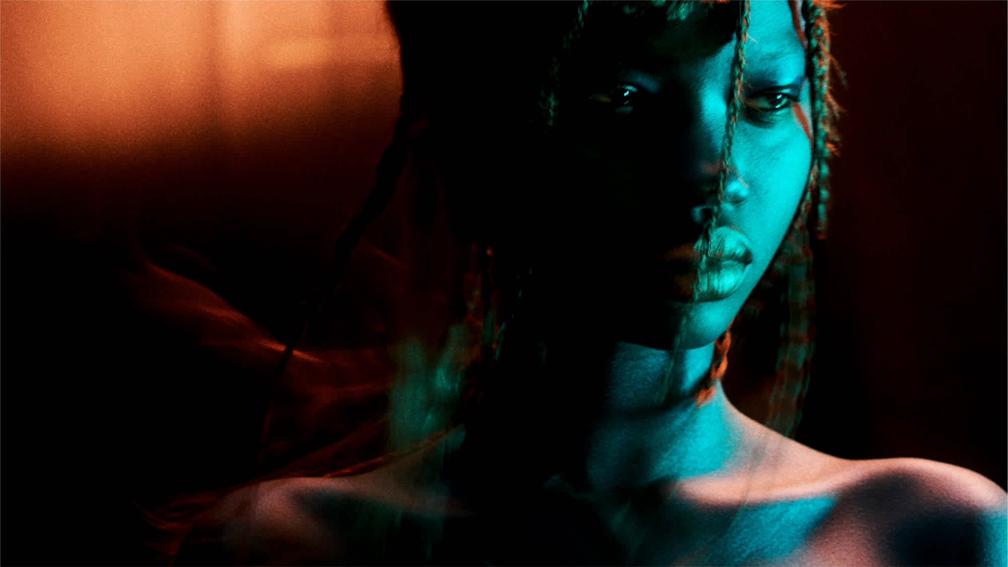 A woman's face is partially illuminated by light blue toned light, and the other half in shadow. The model has braids, and shoulders exposed. There is a warm toned light in the upper left hand corner of the frame.
