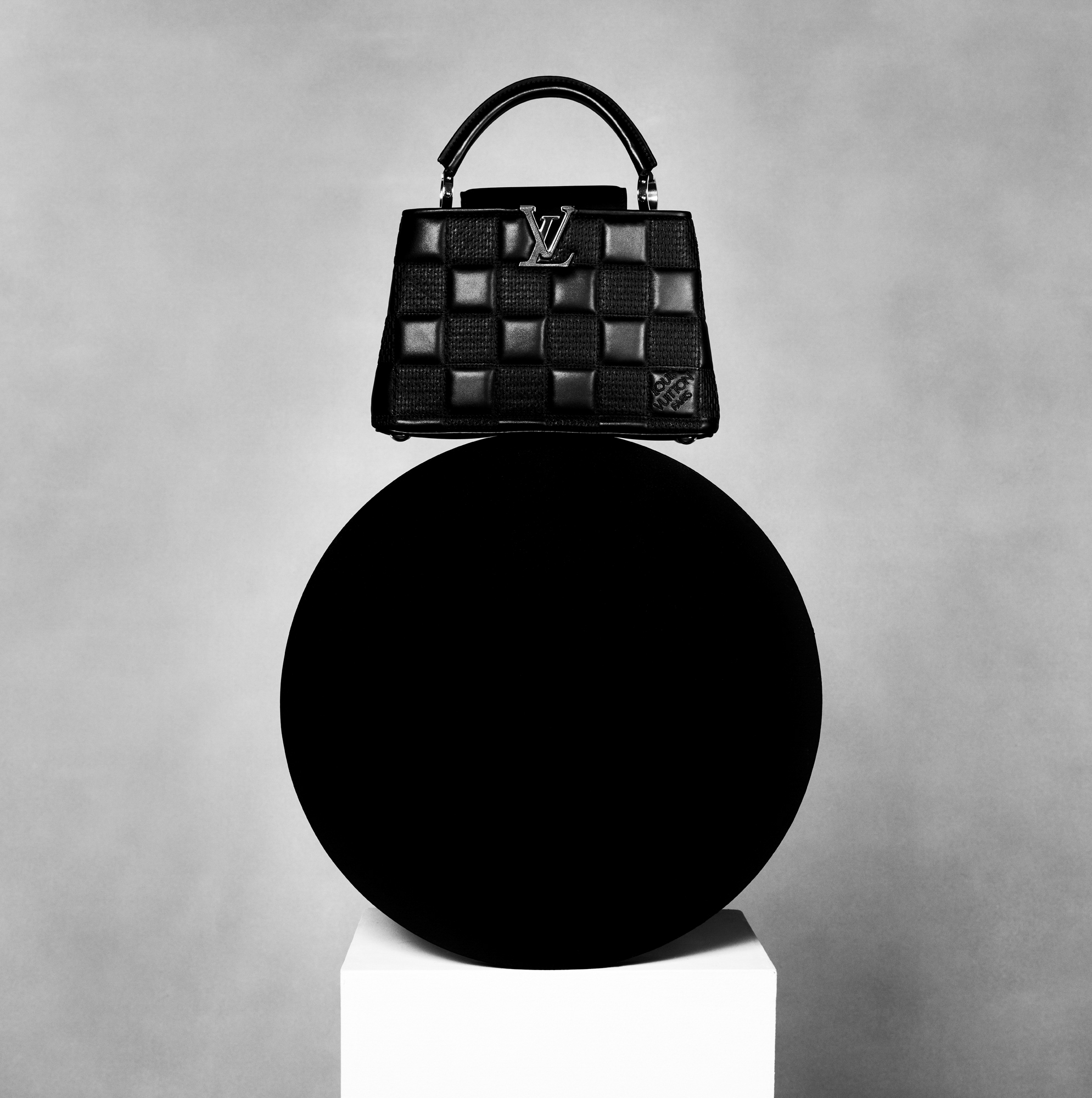 A small black quilted leather Louis Vuitton bag is resting atop of a black sphere that is supported by a white block. The background is a textured grey. 