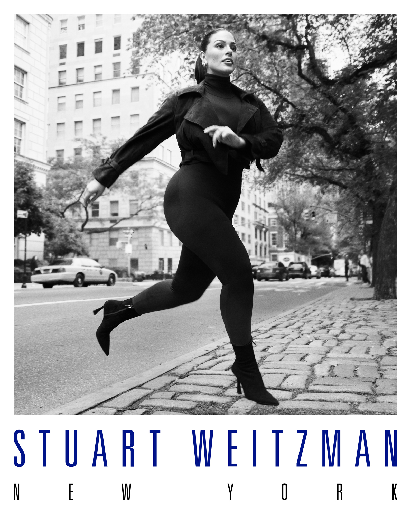 Ashley Graham is wearing a fitted black athletic long sleeve and leggings. She is wearing a small black heel. The image captures her mid stride coming onto the sidewalk crossing a New York City street. The Stuart Weitzman New York is featured at the bottom of the image. 