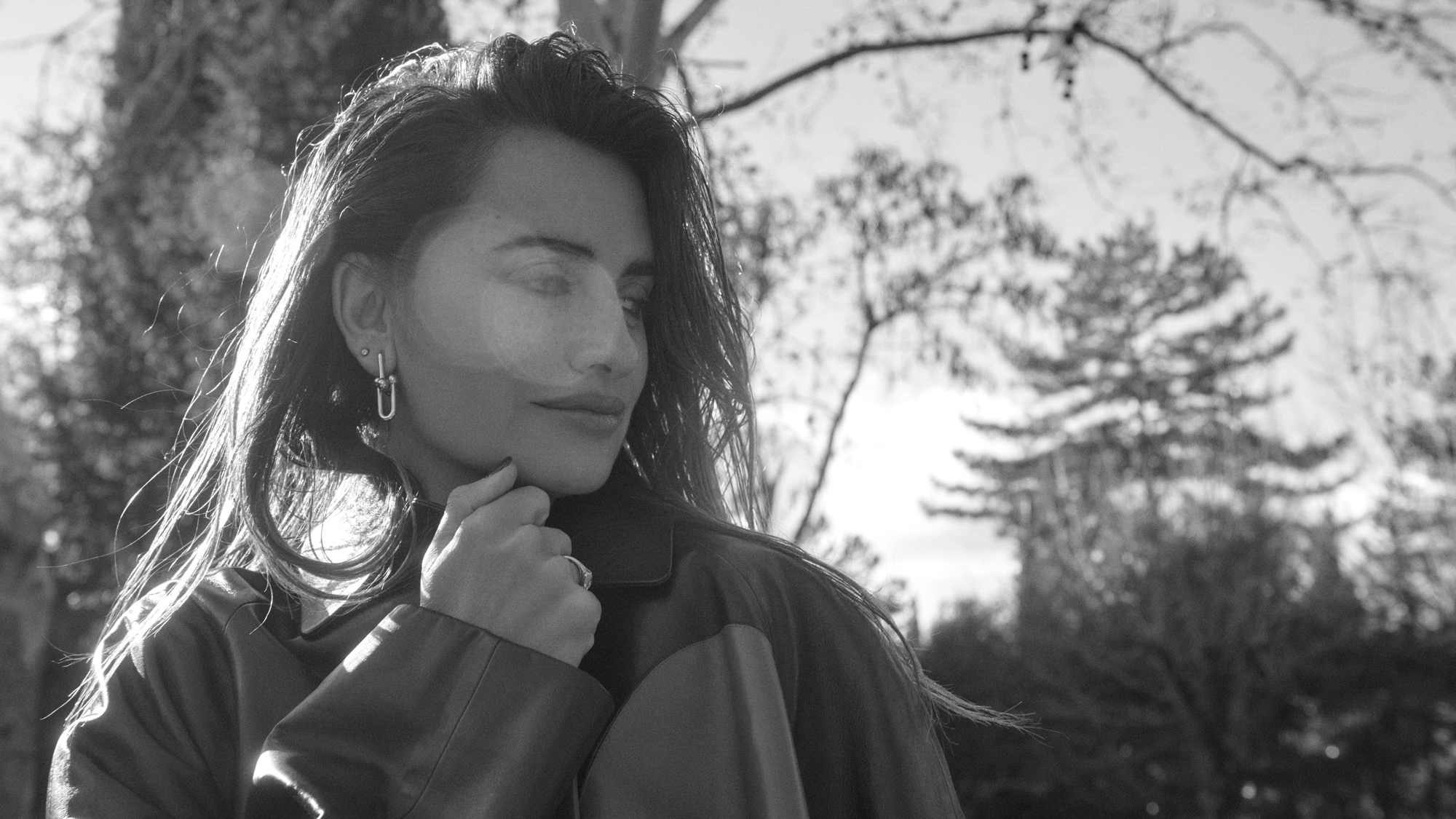 Portrait of Penelope Cruz wearing a thick jacket outside with trees in the background. She is pulling her collar up higher with one of her hands. 