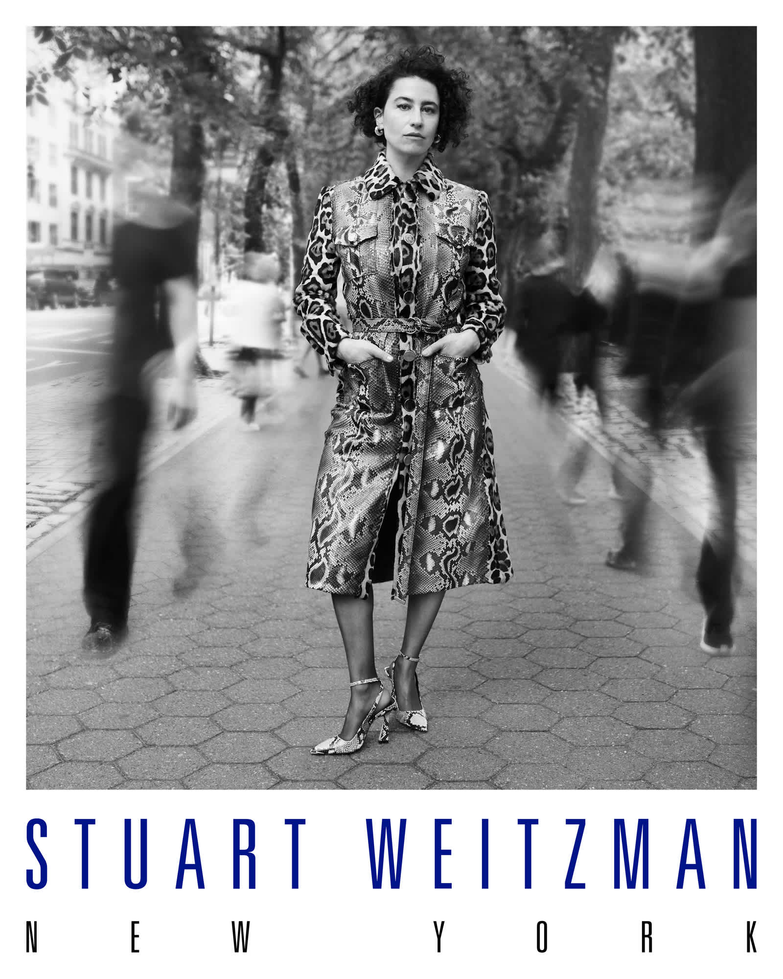 Ilana Glazer is wearing an animal print knee length jacket with heels on. She has her hands in the pockets of the jacket and is looking directly at the camera. People are motion blurred around her as they walk down the sidewalk. The Stuart Weitzman New York logo is featured directly below the image.