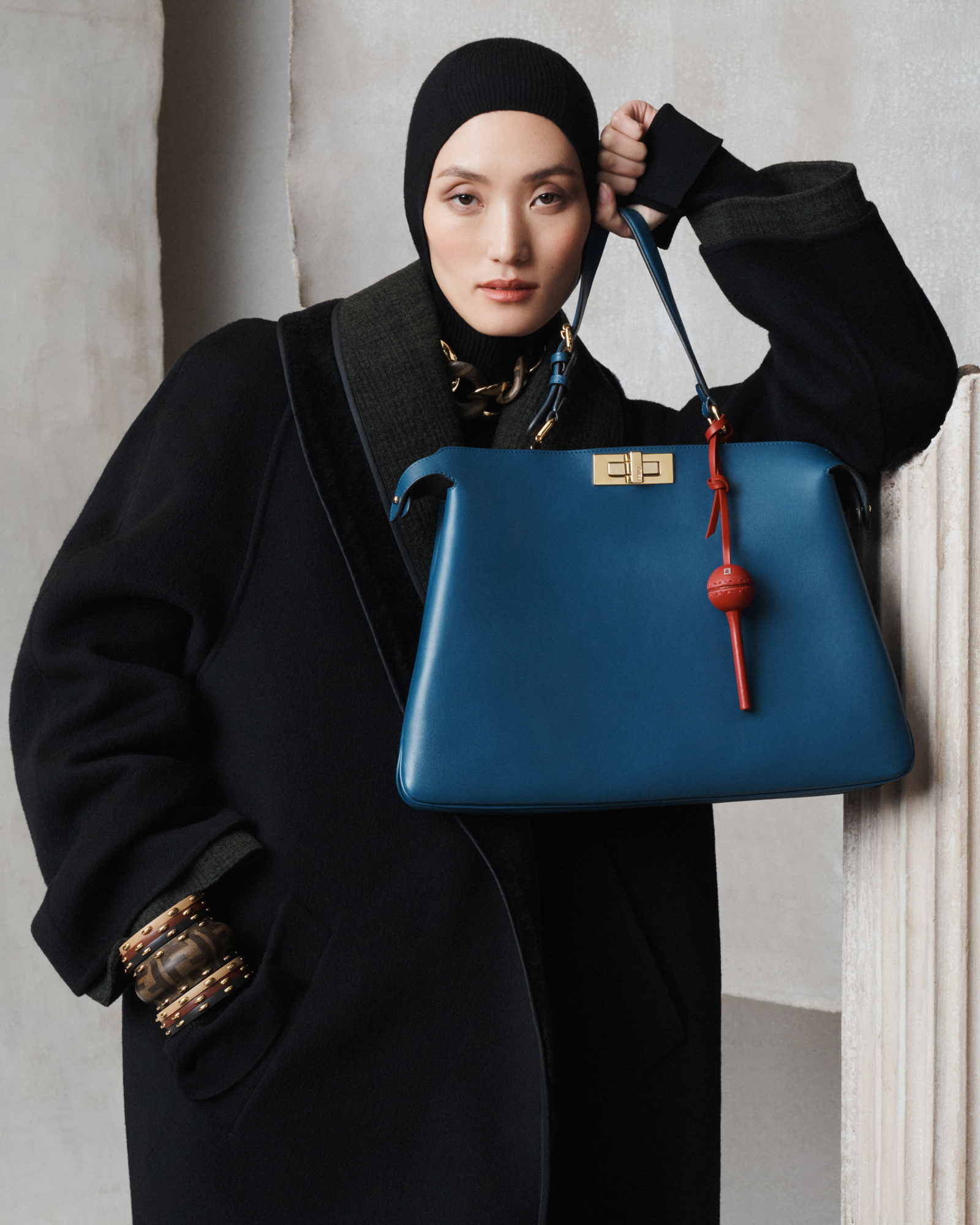The model is wearing a thick dark coat with a black head covering. She is leaning on elbow on the top of a Greek column, and holding a blue leather medium sized Fendi bag in one hand. Her other hand is in her pocket, highlighting the brown toned bracelets on her wrist. Different pieces of textured and smooth gray papers are in the background. 