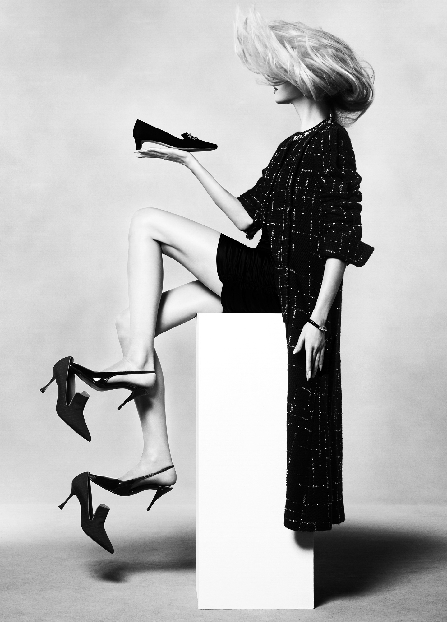 The model is seated on a white column. She is in profile holding on shoe in her hand, and balance two other shoes on both feet. Her hair is being blown forward, obscuring her face. 
