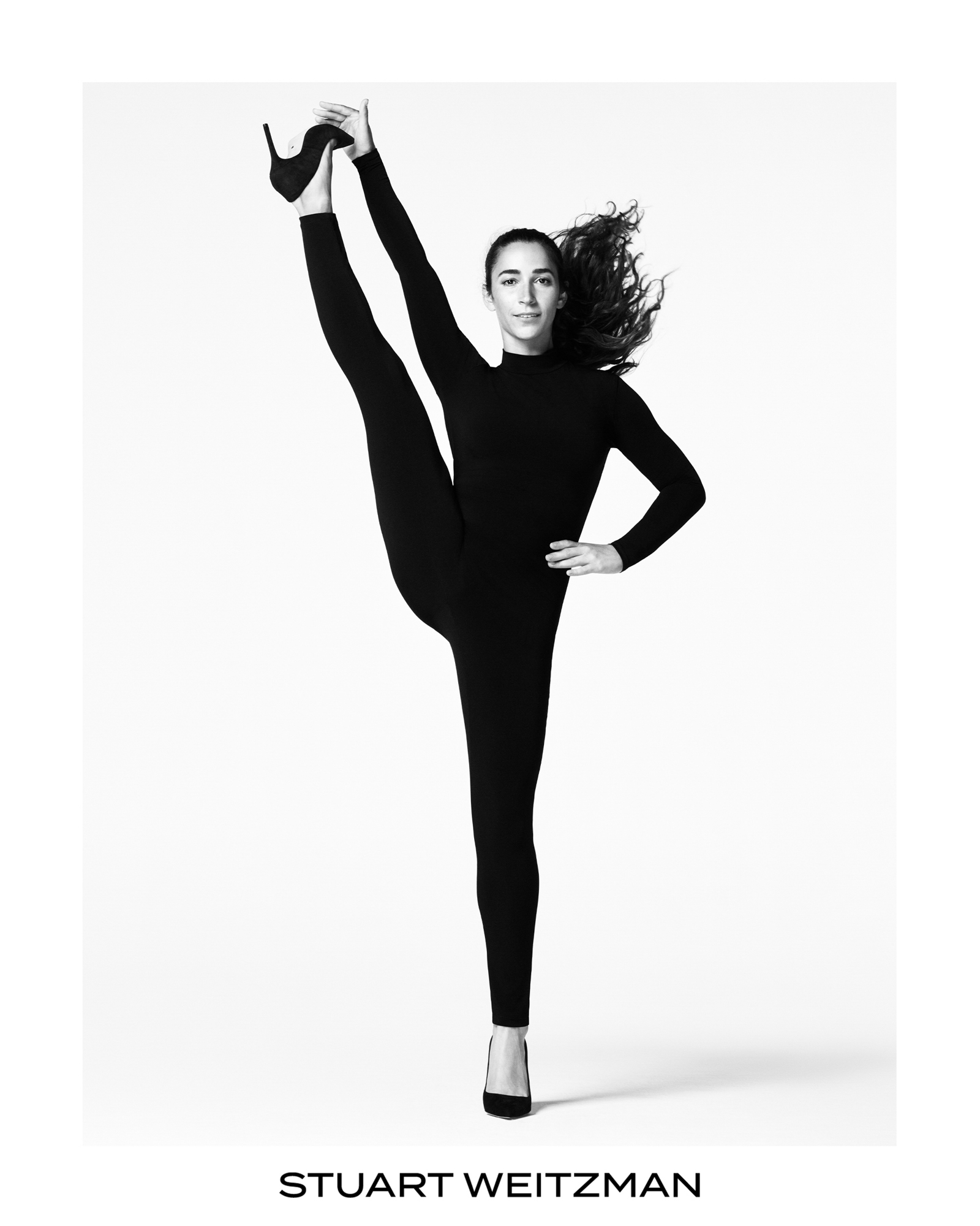 Aly Raisman has one leg fully kicked up into the air, she is holding the bottom of her foot that is raised in the air. She is wearing an all black form fitting body suit, she has simple black pumps on. She is facing the camera with one hand of her hip. Stuart Weitzman logo is featured on the bottom center of the frame. 