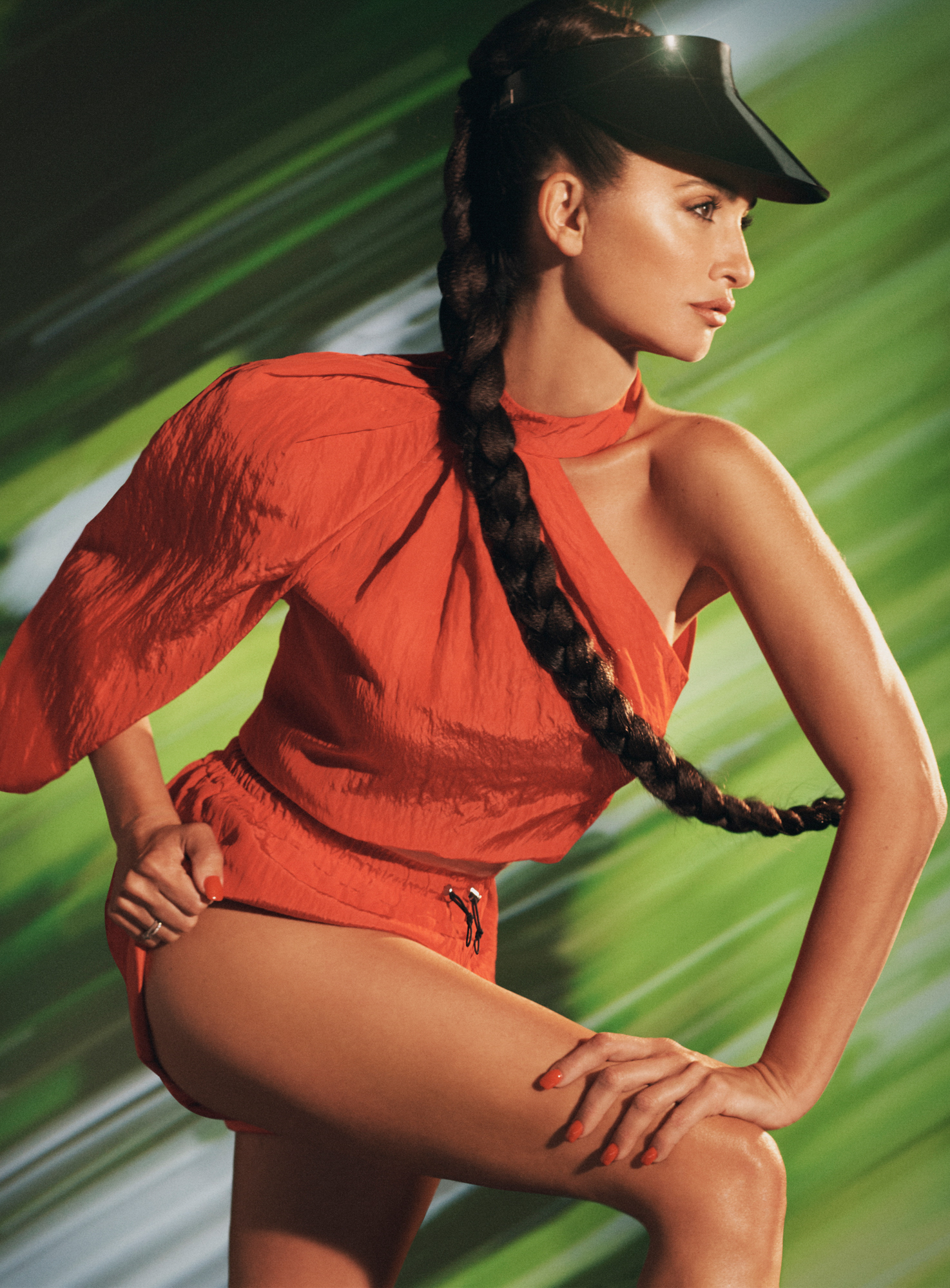 Penelope Cruz is leaning forward in front of a green background. She is wearing an orange top with orange shorts, has a black visor on, and a long braid. 