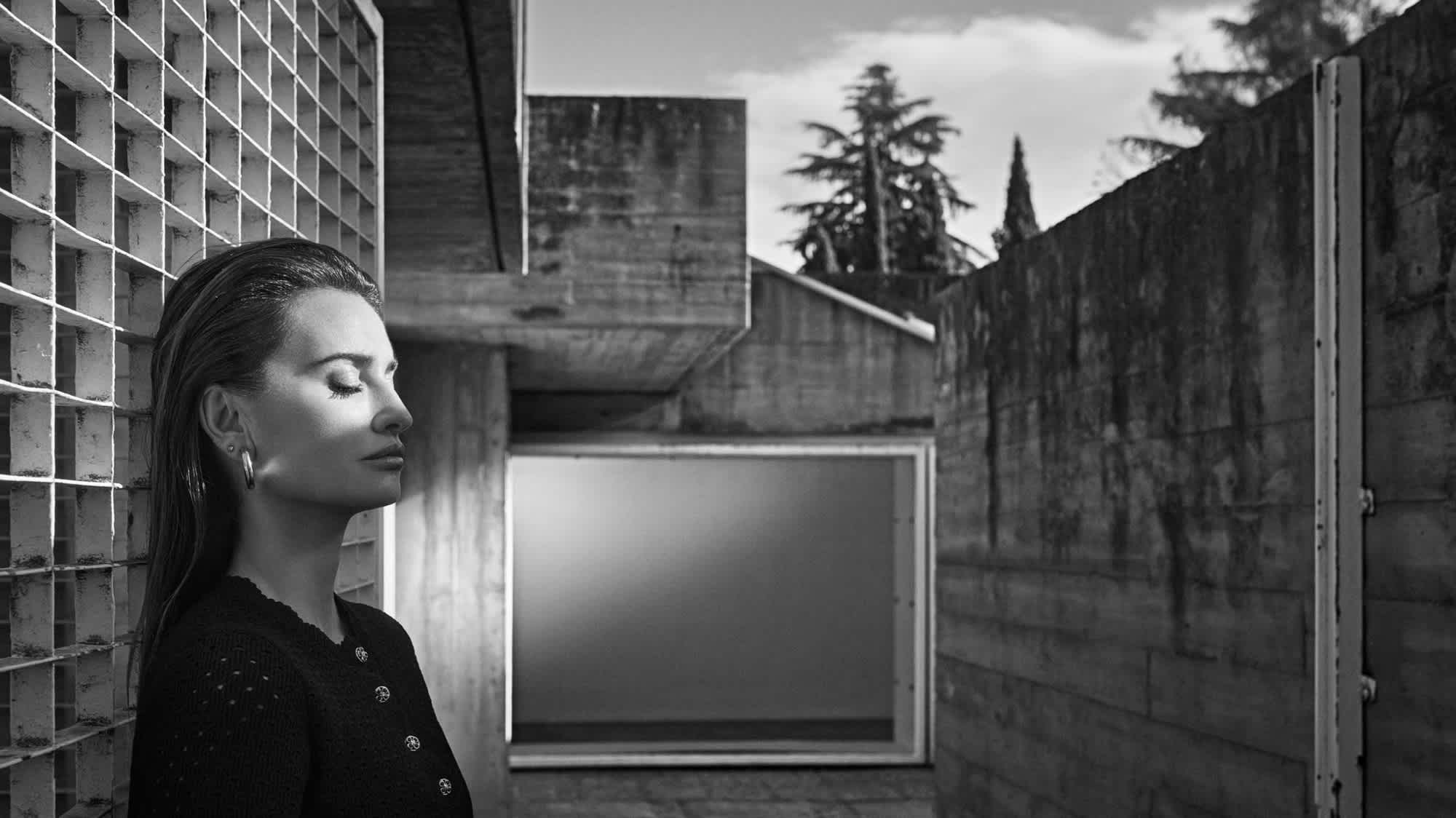 Penelope Cruz is standing with her eyes closed against a wall with grid like openings. She has light hitting the top half of her face. She is standing in what appears to be some kind of courtyard with more Brutalist architecture.