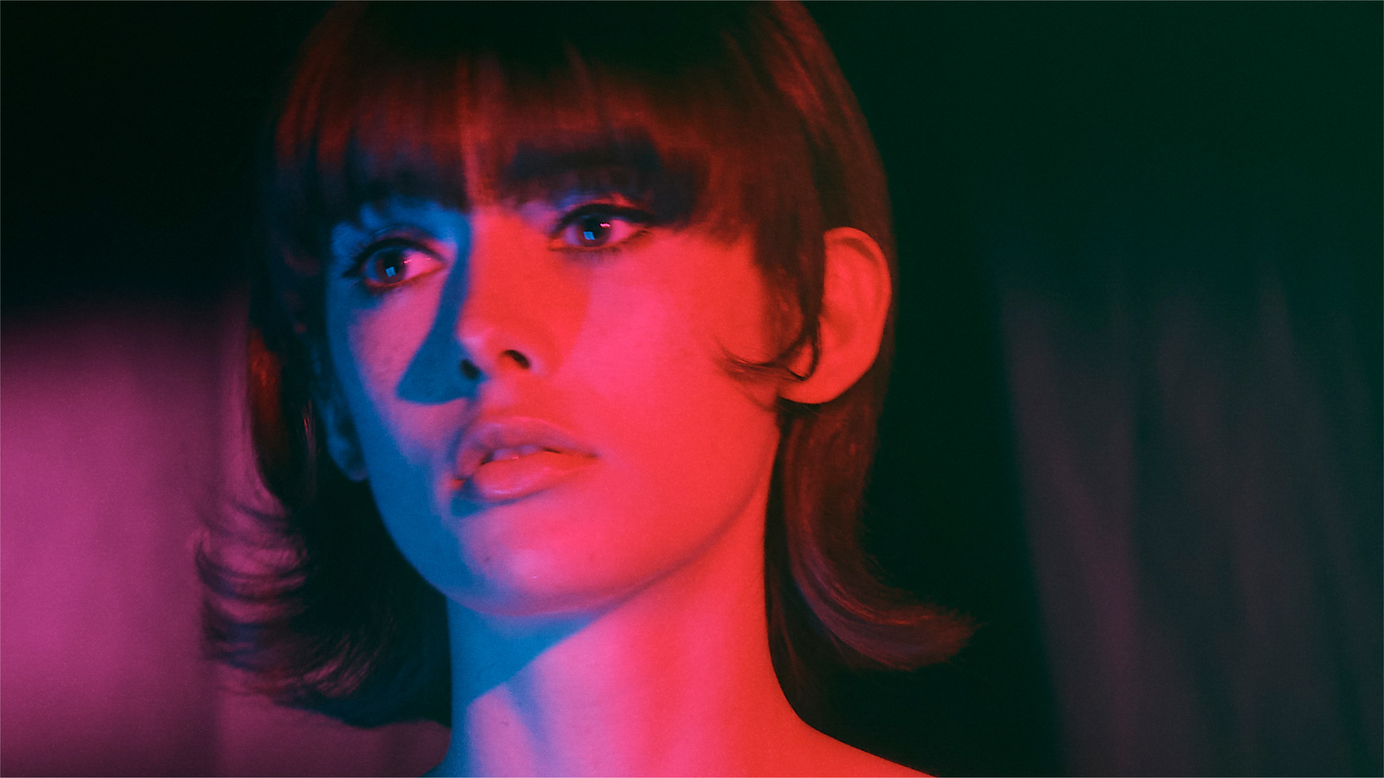 A woman with a 60s Mod haircut is looking off camera to the left side of the frame. She has graphic black eyeliner, and her face is being lit by both blue and red light. The background is a fabric that is being hit with purple toned light. 
