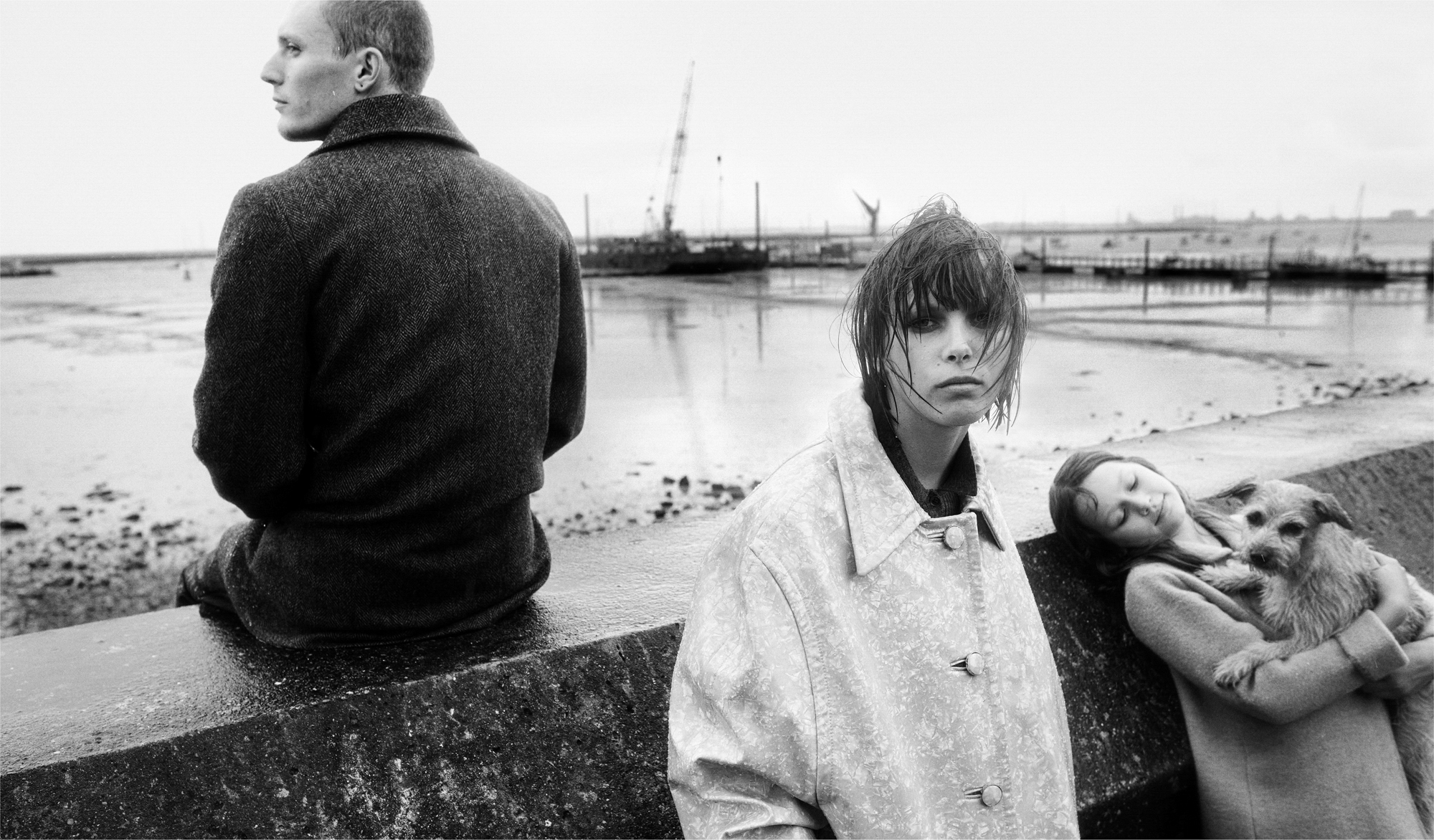 A woman with wet hair in a coat is looking at the camera while a young girl with a dog is leaning against a stone wall. A young man is seated on the stone wall in a coat looking off to the left side of the frame. 