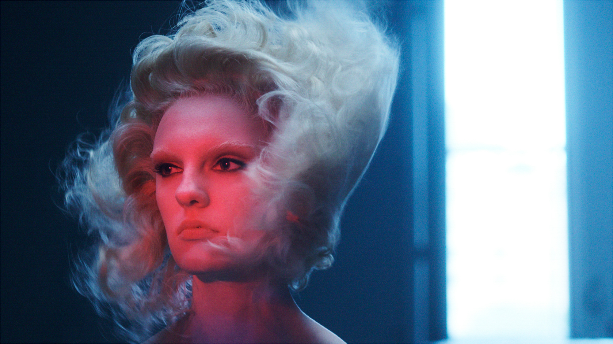 A woman with blonde wavy hair is getting blown by wind towards the back of her head. Hair is blowing around the face. Her face is being hit with a spotlight of red toned light, she has black eyeliner around the eyes. There is a strip of bright light coming from the right side of the frame. 